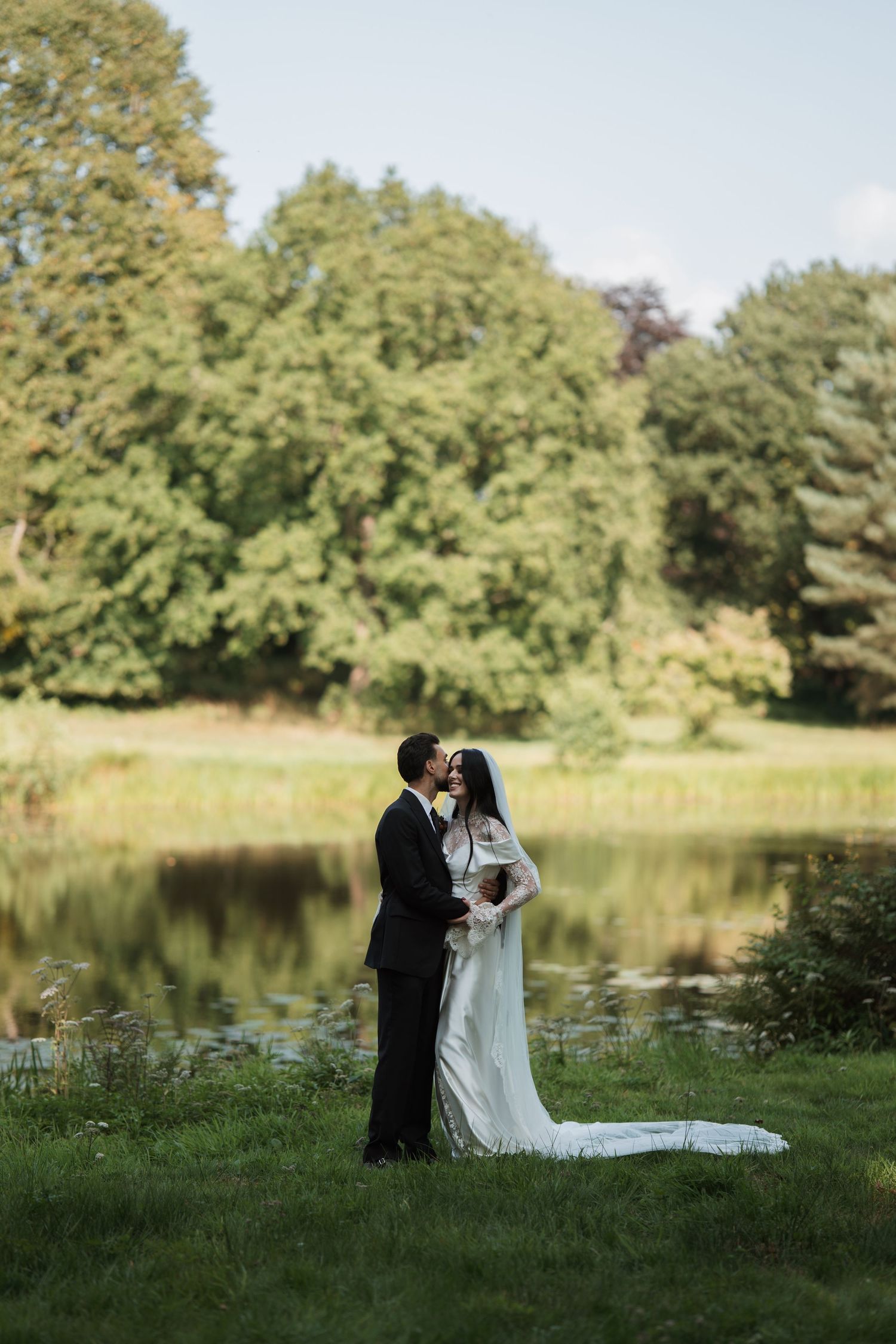 Newlyweds stand by a serene pond surrounded by lush trees in their wedding attire.