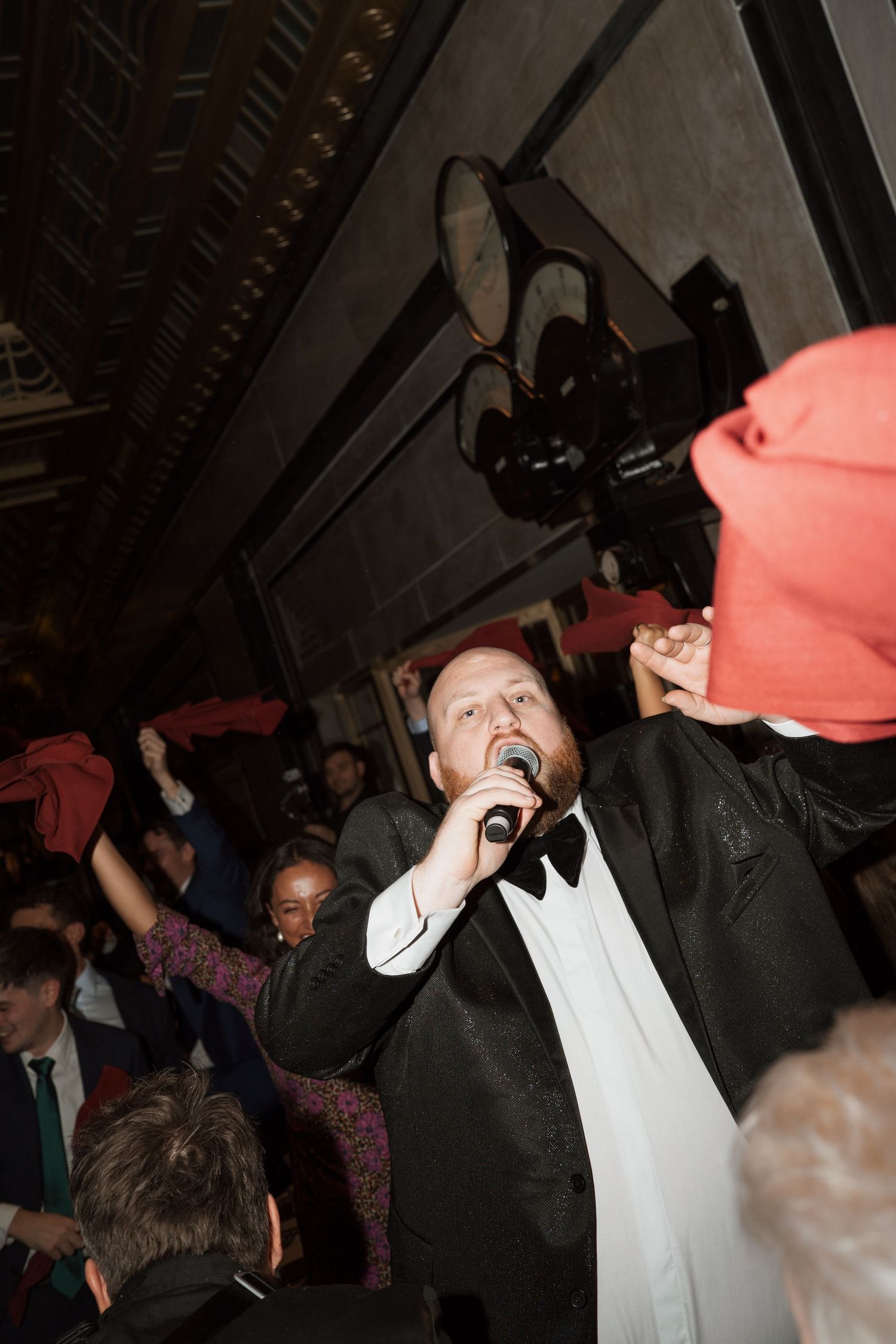 Man in tuxedo singing passionately at formal event while crowd waves red napkins.