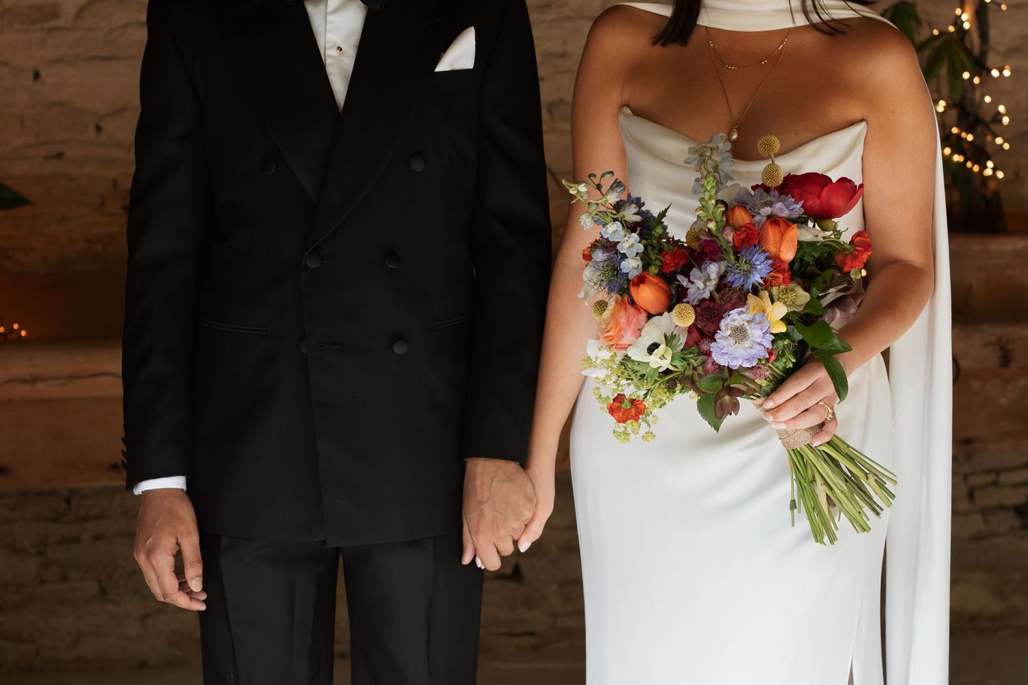 Wedding couple posing with colorful bouquet against stone wall backdrop.