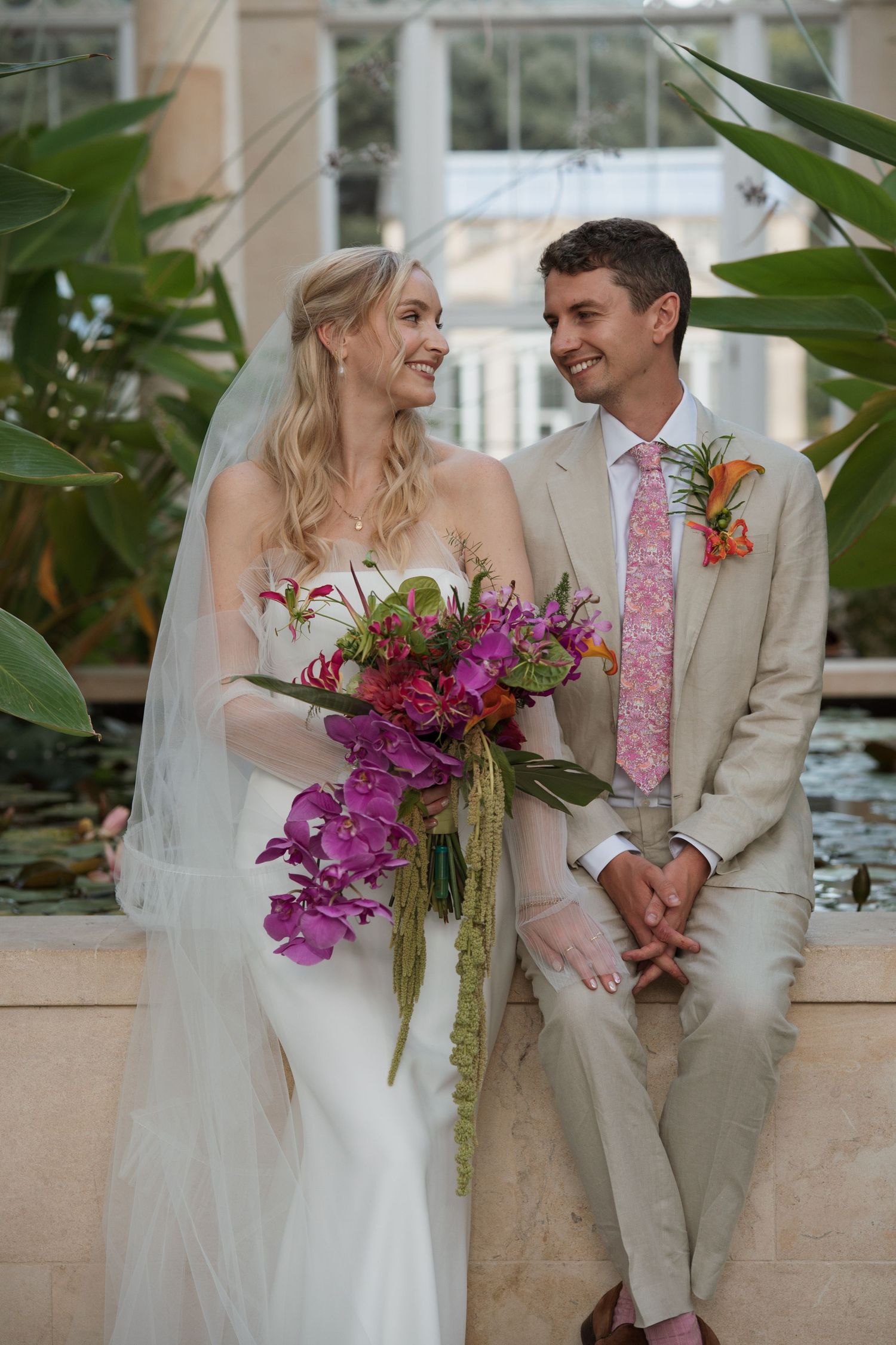 A couple in wedding attire share intimate moments while holding vibrant purple orchid bouquets against a light stone backdrop.