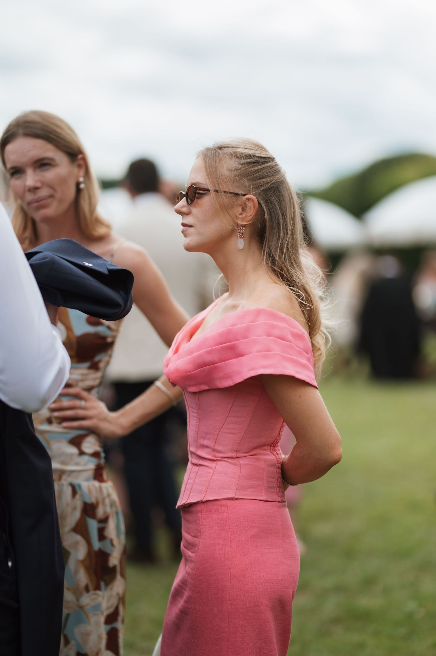 Elegant woman in pink off-shoulder dress and sunglasses at an outdoor garden party event.