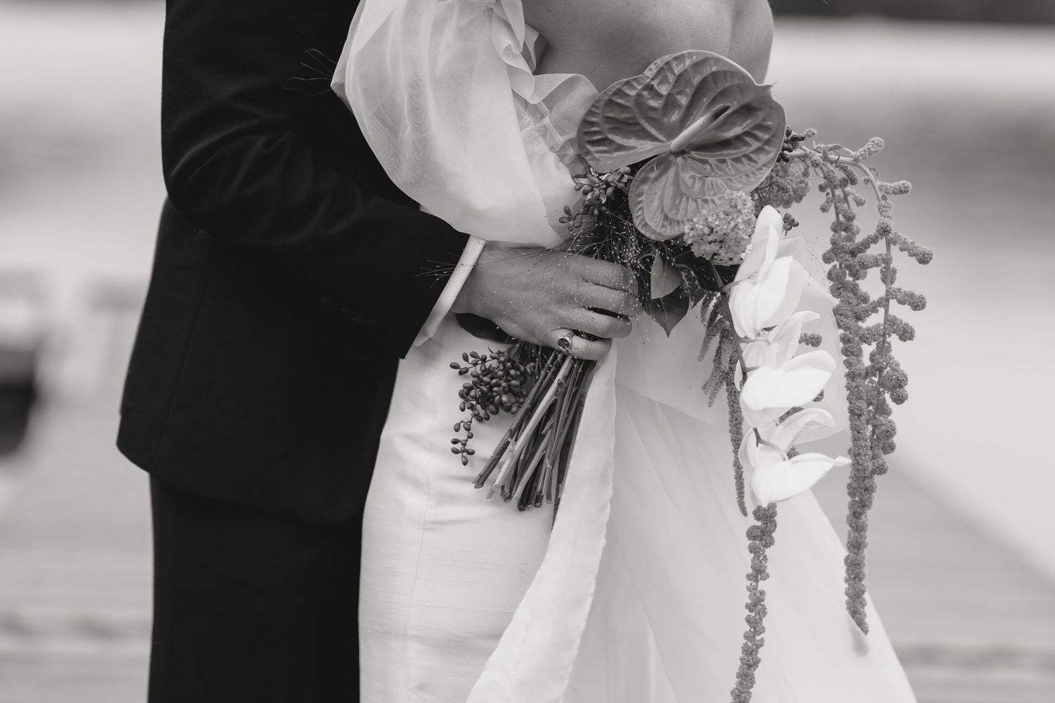 Black and white photo of a bride and groom embracing, bride holding a floral bouquet with trailing greenery.
