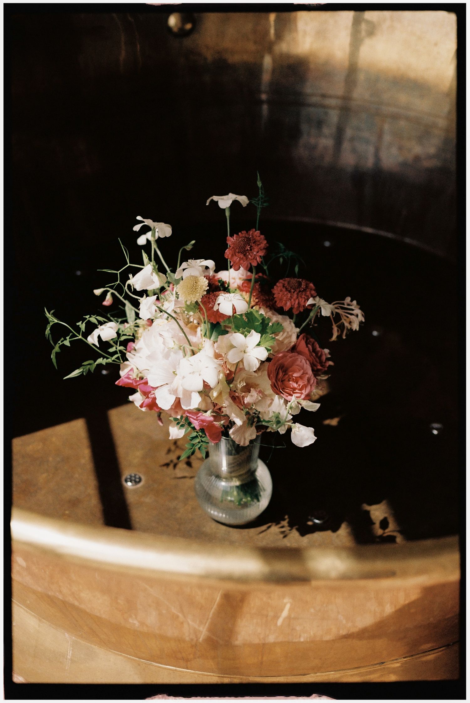 A vintage floral arrangement of white peonies, dusty rose blooms, and greenery in a small silver vase on a wooden barrel.