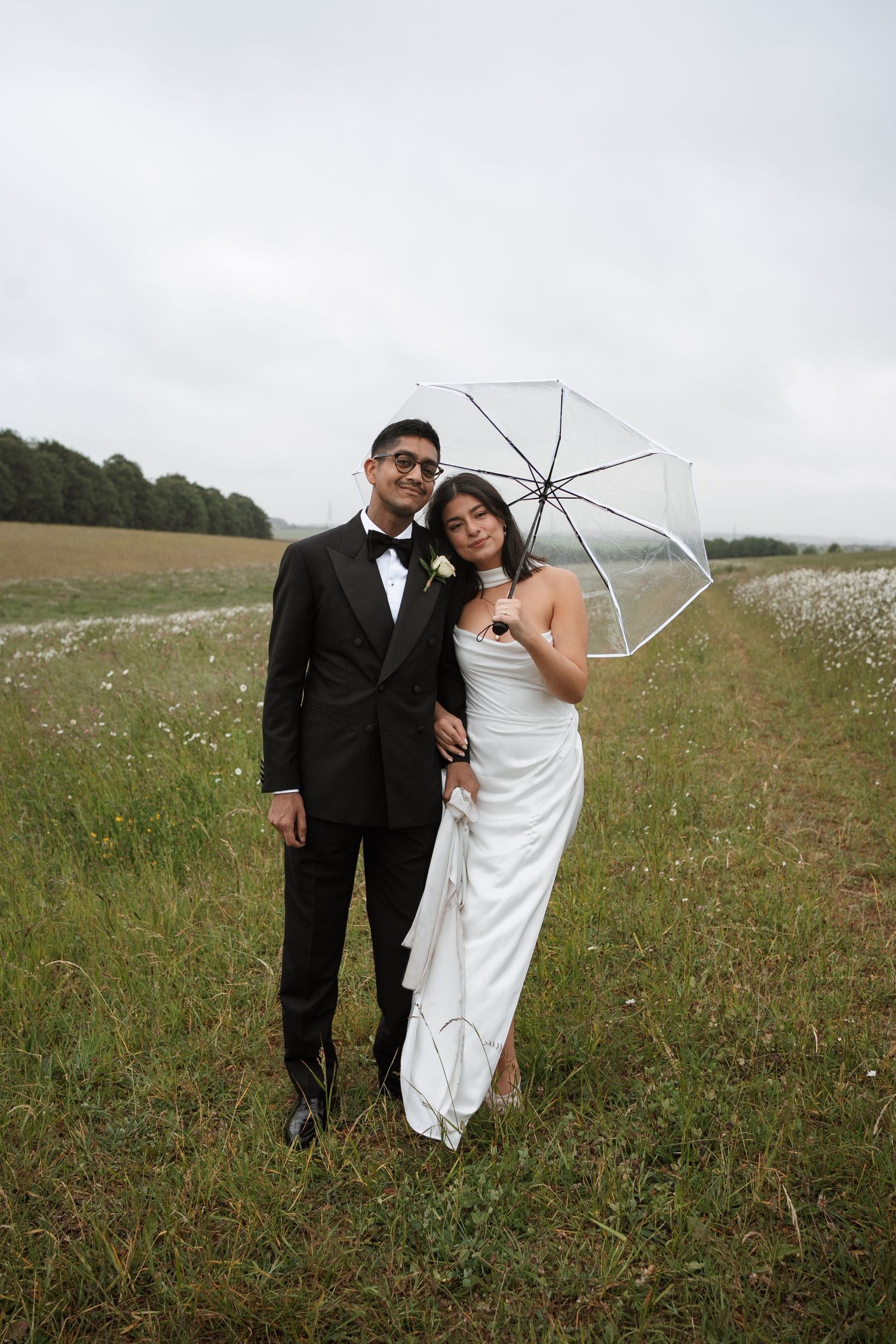 A couple walks through a wildflower field holding a clear umbrella on their wedding day.