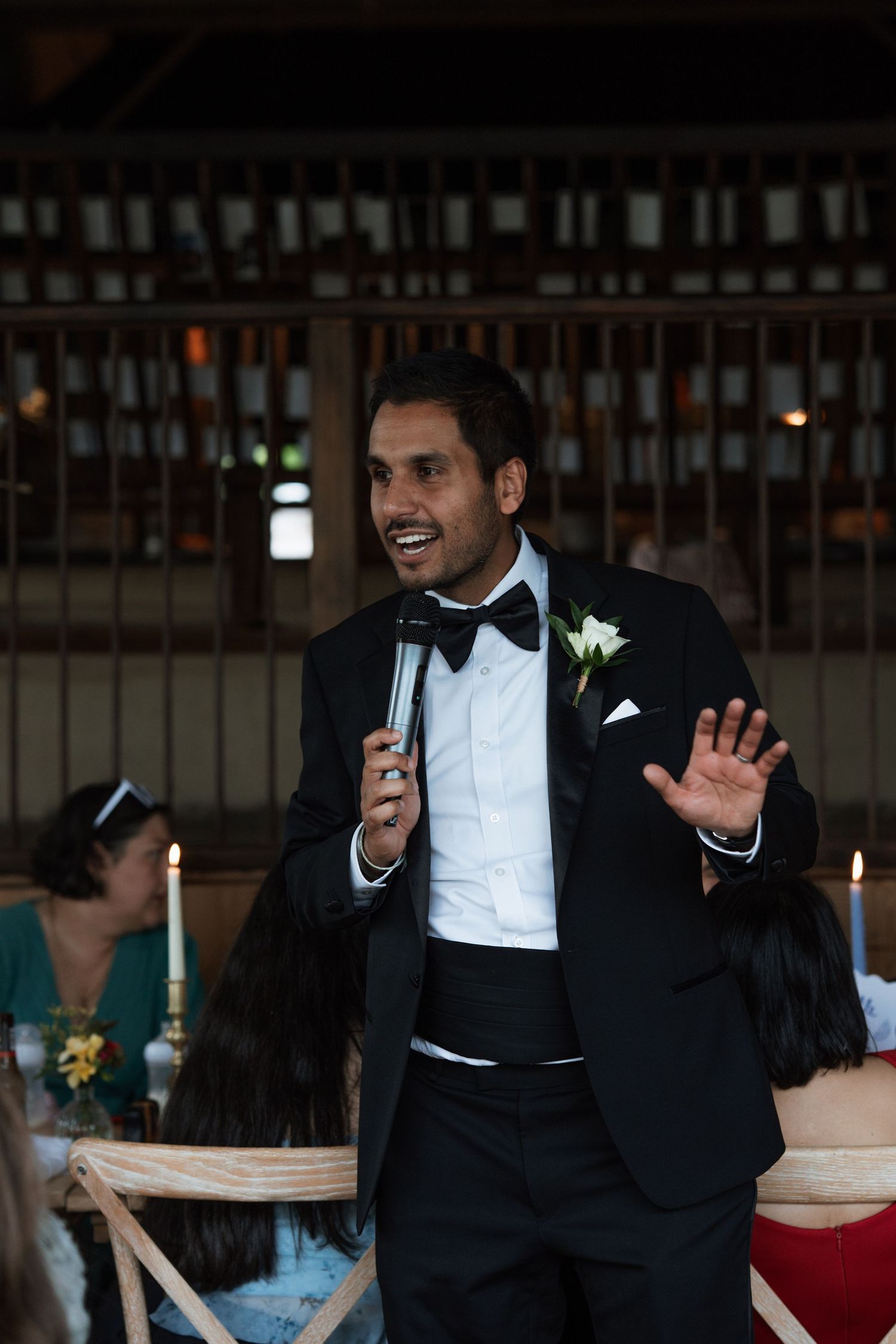 Speaker in formal black tuxedo giving an animated wedding toast at an indoor reception.