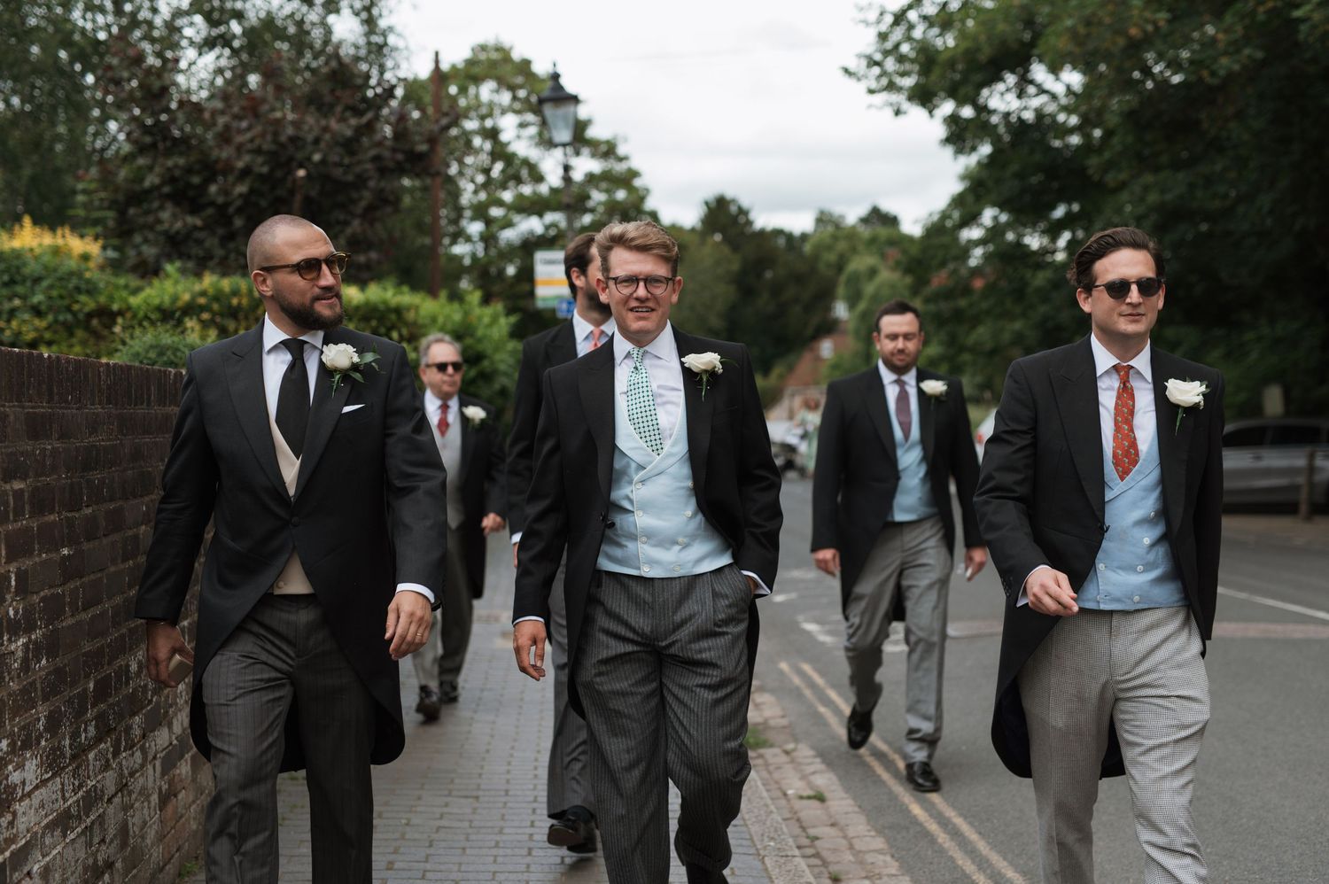 Groomsmen in morning suits walking down a street on a wedding day, led by the groom in a light blue tie.