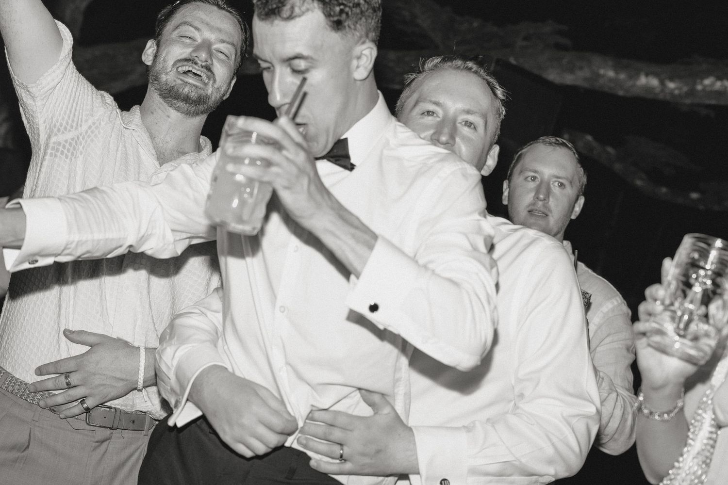 Men in white dress shirts celebrating at a wedding reception party, drinking and dancing together in black and white photo.