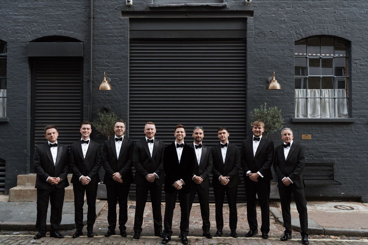 Nine groomsmen in black tuxedos and bow ties posing in a row outside a dark brick building.