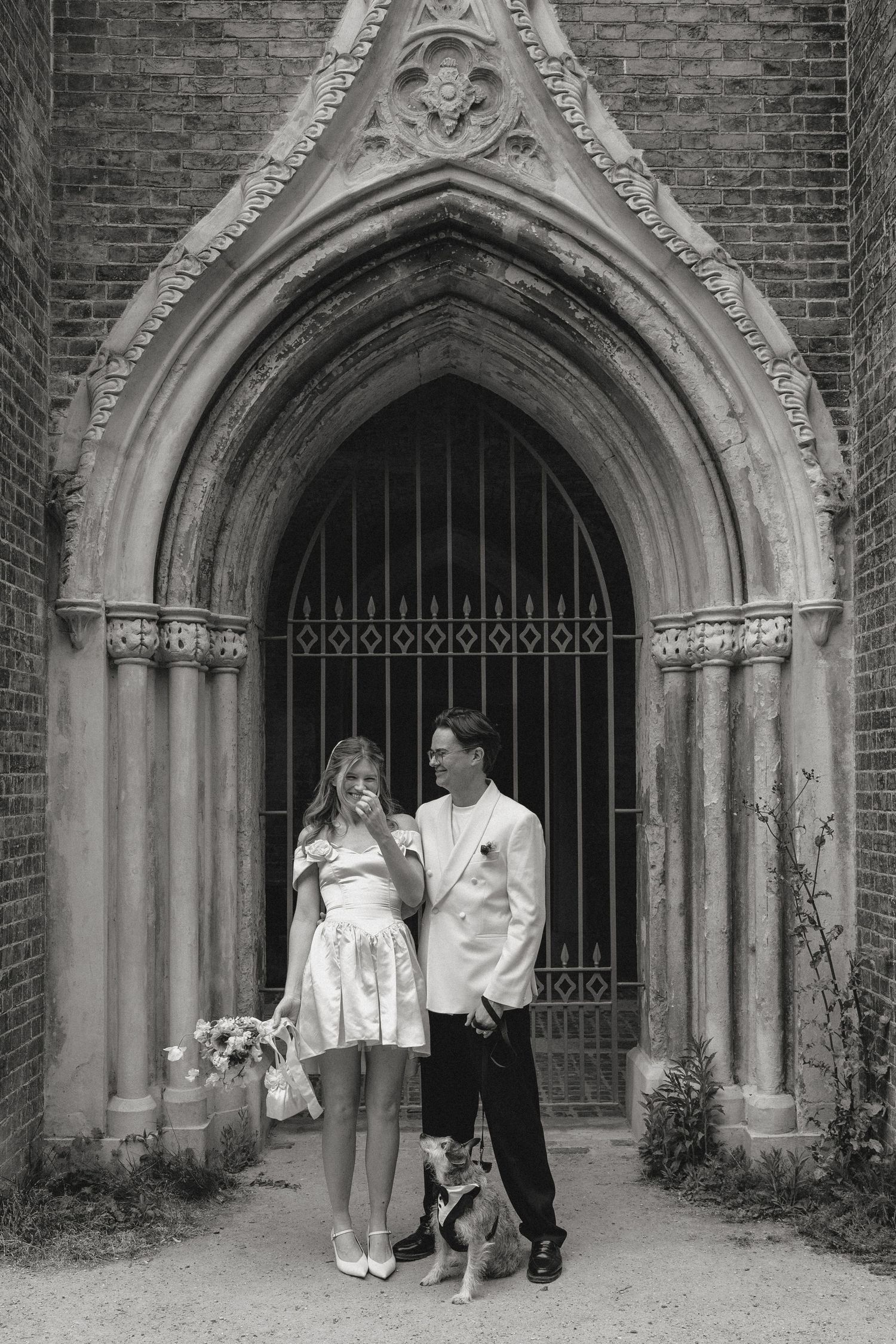 Black and white photo of a couple in wedding attire standing before a Gothic church arch with a small dog.