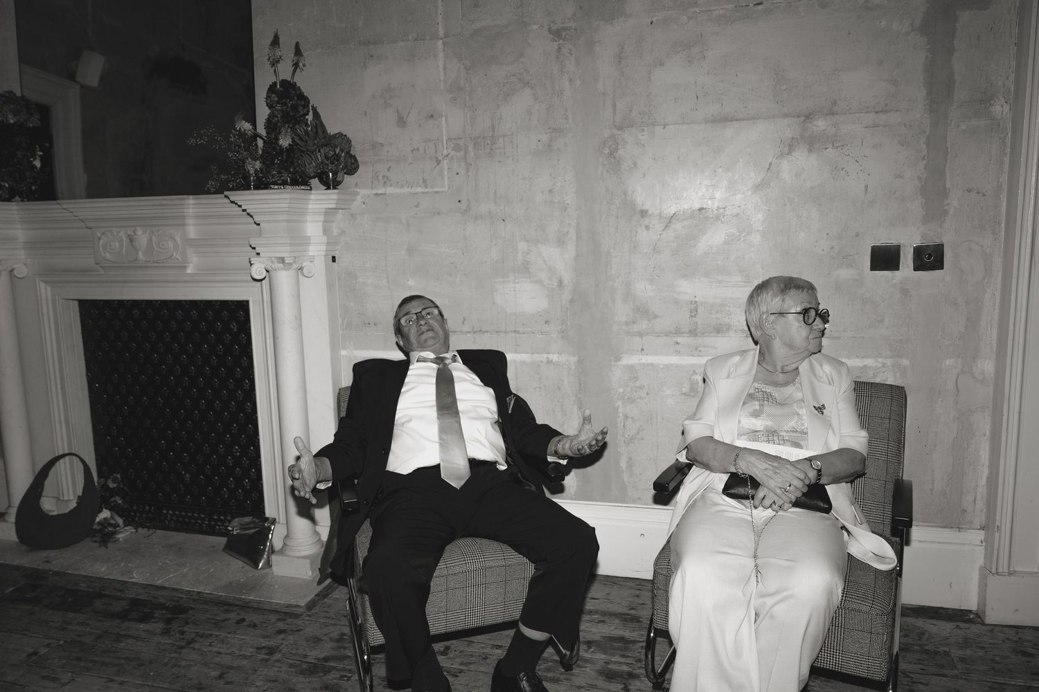 Black and white photo of a man in a suit dozing in a chair beside a smiling elderly woman near a stone fireplace.