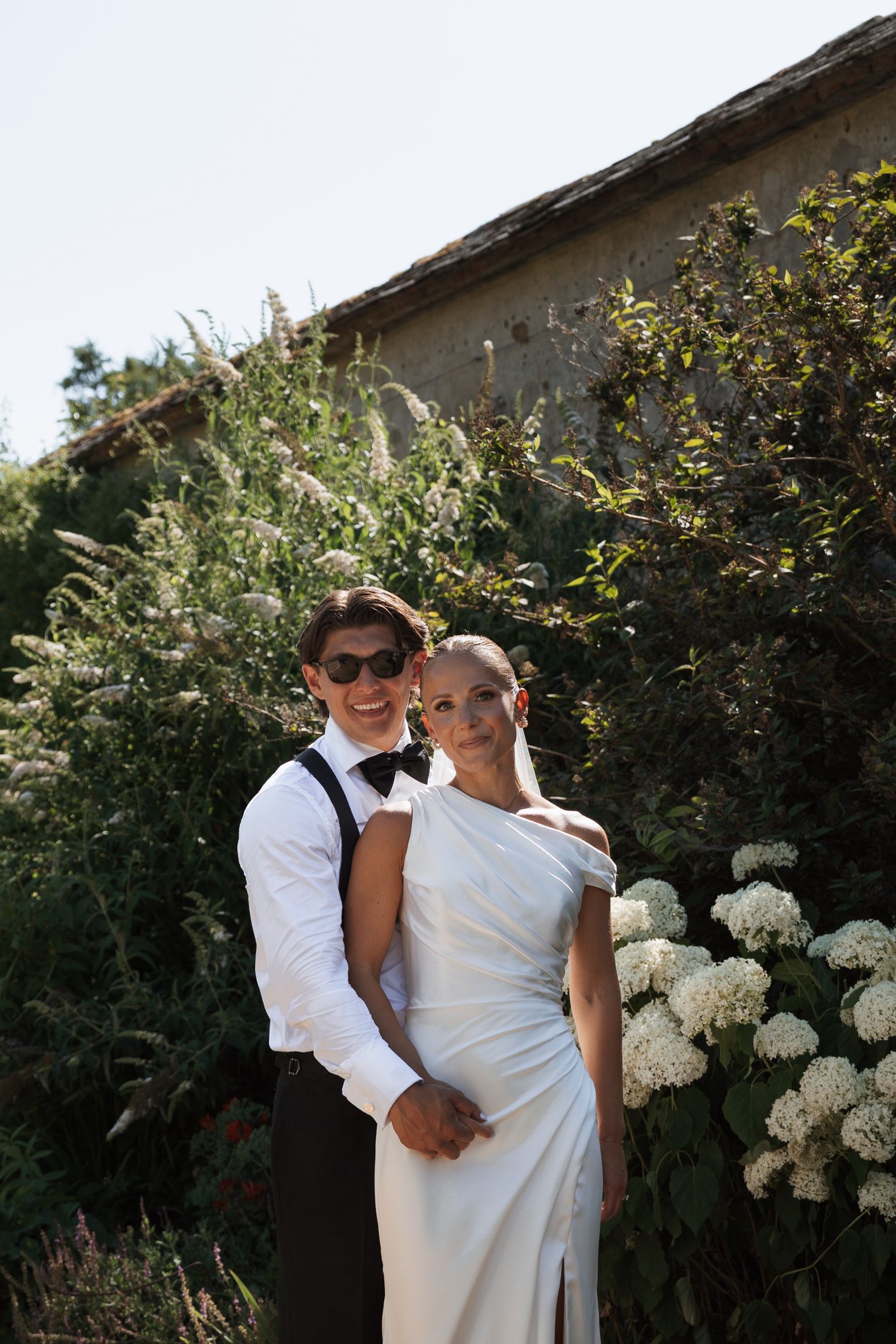 Elegant bride in white gown and groom in suspenders pose together in lush garden with hydrangeas.