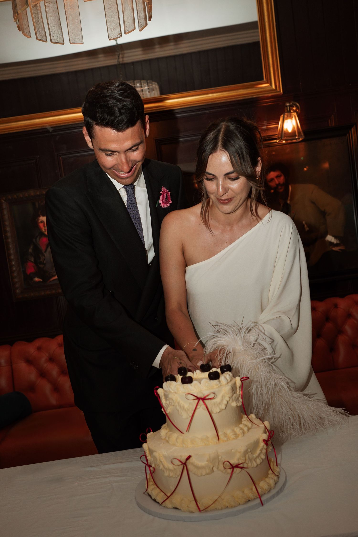 Wedding couple cutting into an elegant white cake decorated with feathers in a dimly lit reception venue.