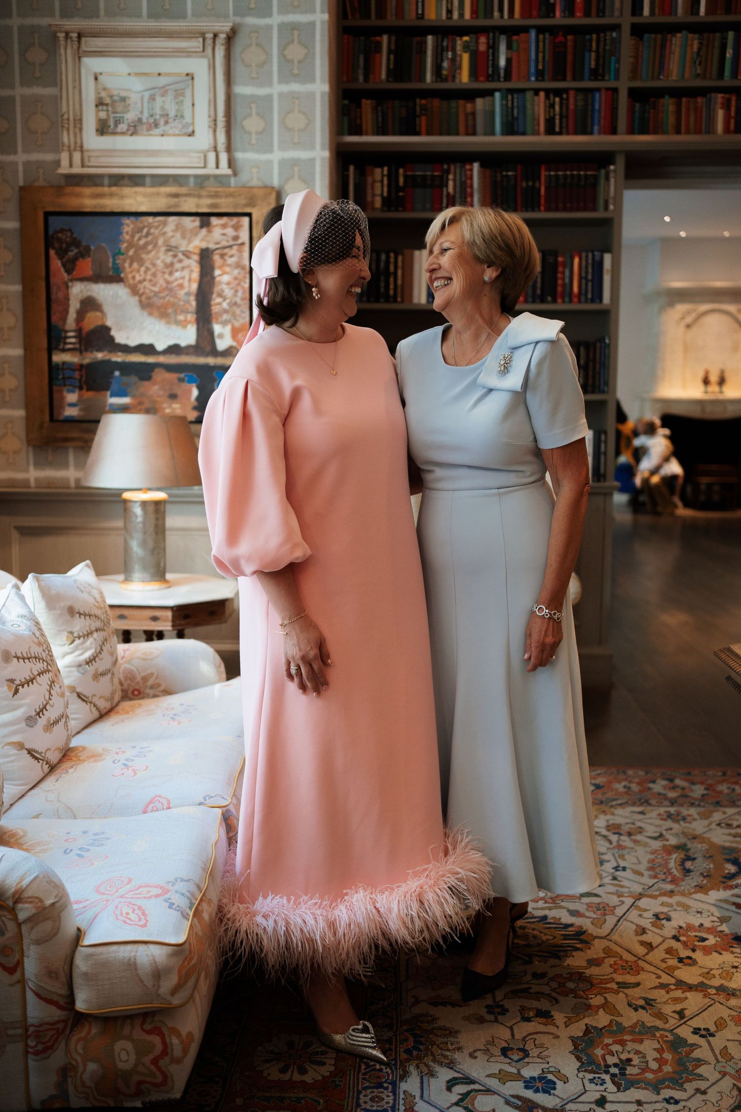 Two women share a joyful moment in an elegant library, wearing formal dresses in pink and blue with feather trim.