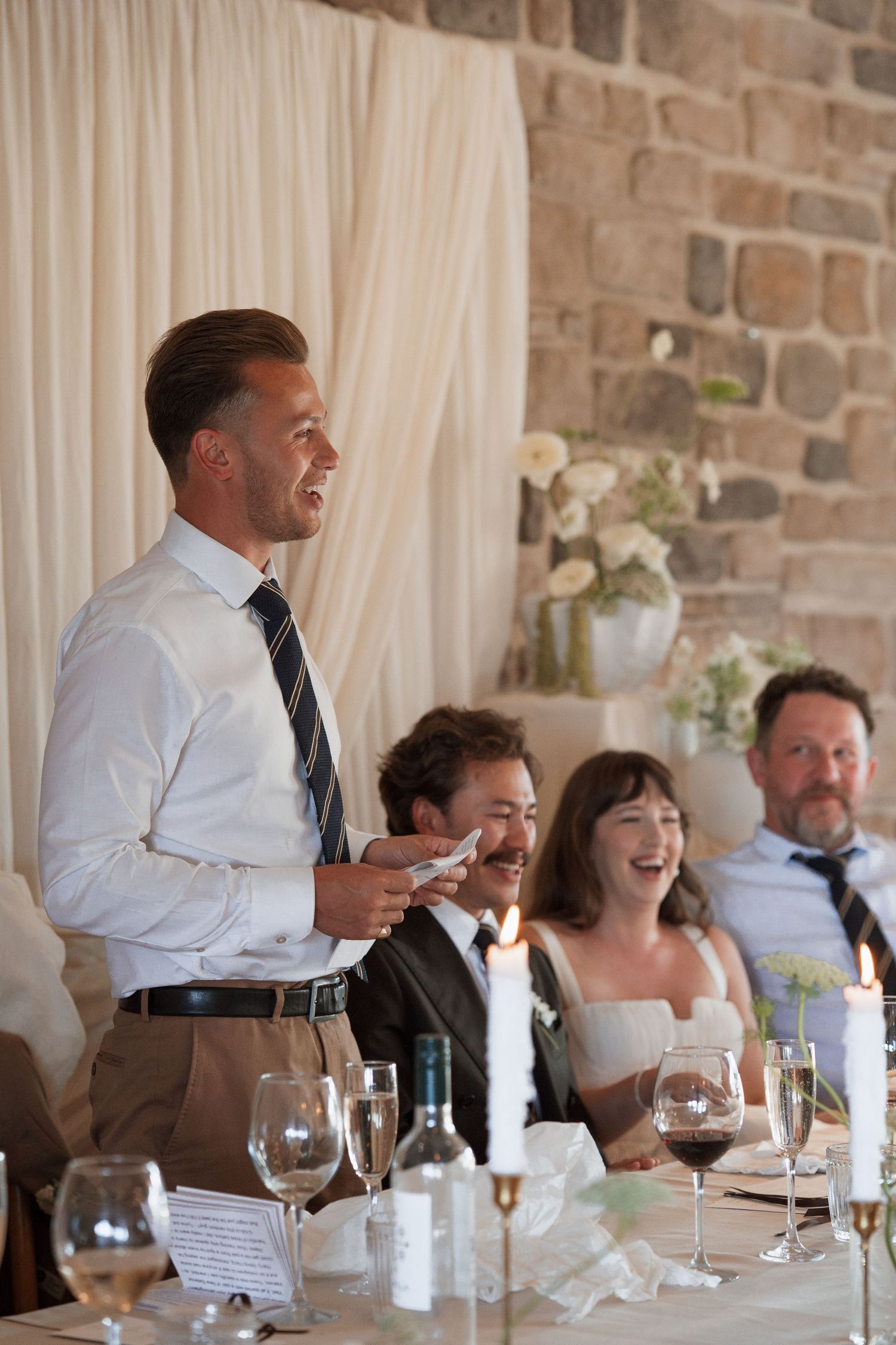Wedding guests sharing joyful moments during reception dinner against stone wall backdrop.