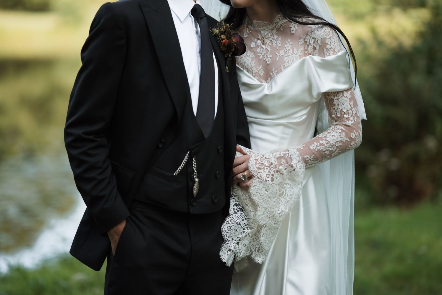 A couple in wedding attire stands close together near a pond in an outdoor setting.