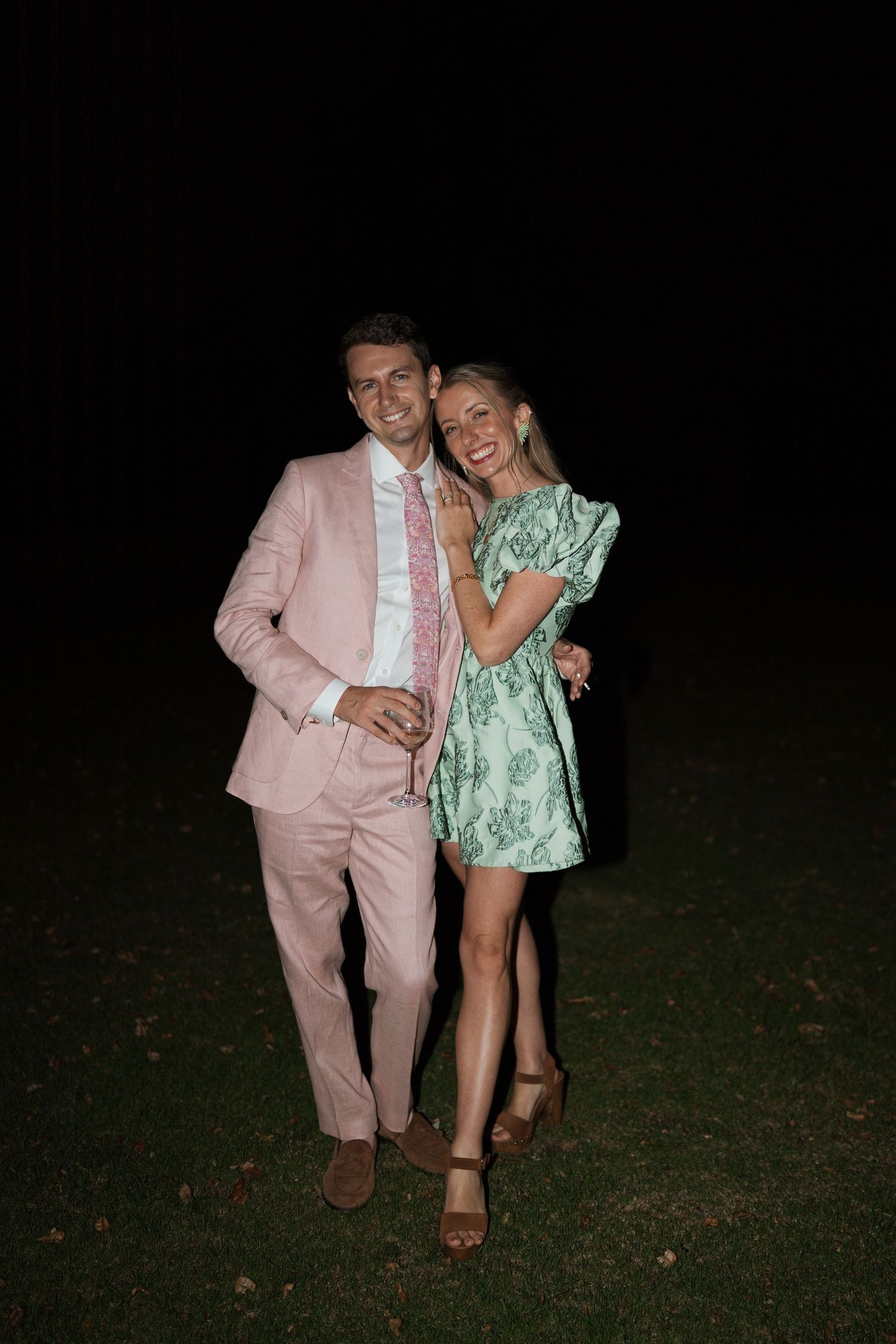 Couple in pink suit and green dress posing together outdoors at night.