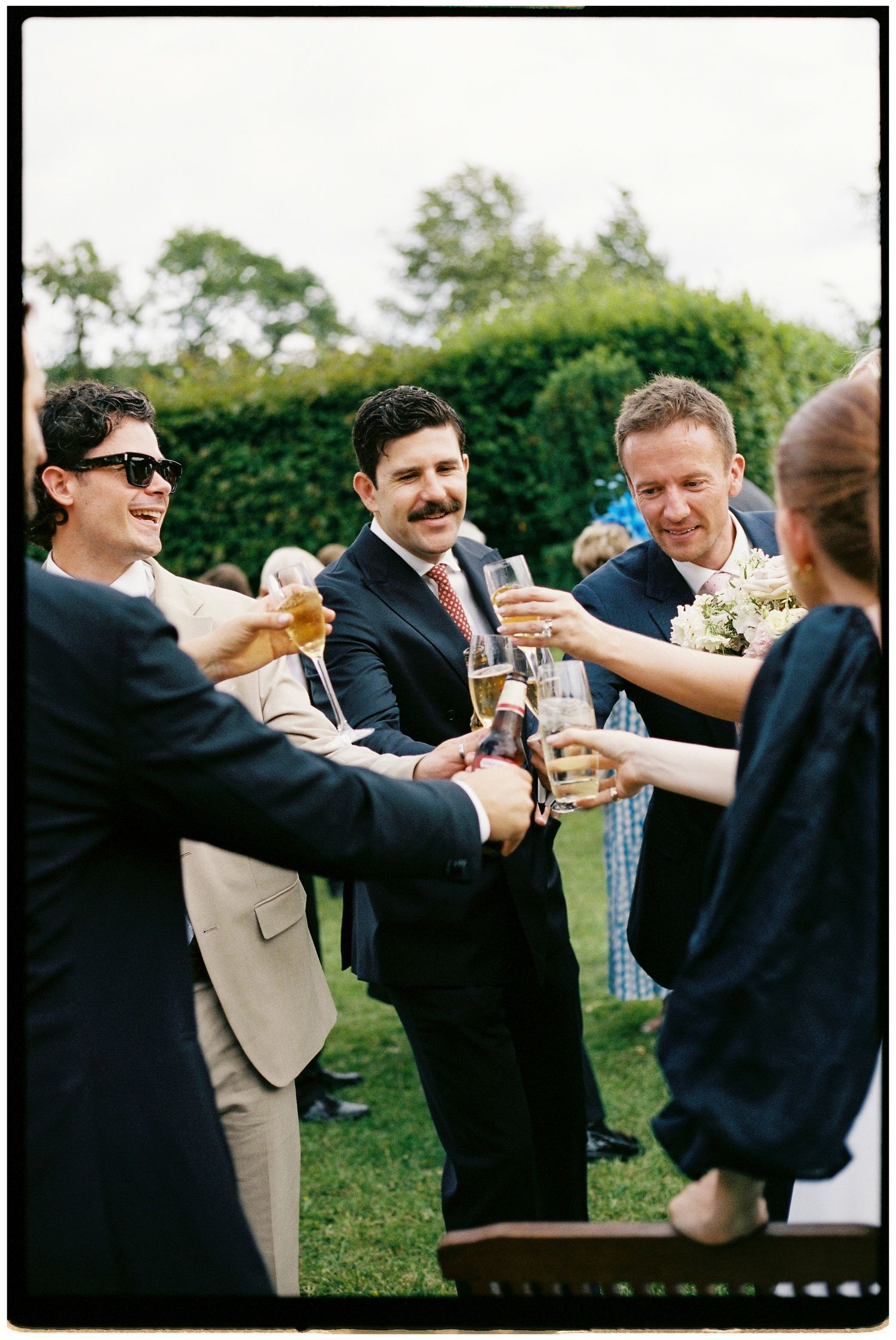 Group of wedding guests in suits toasting with champagne glasses outdoors at a garden reception.