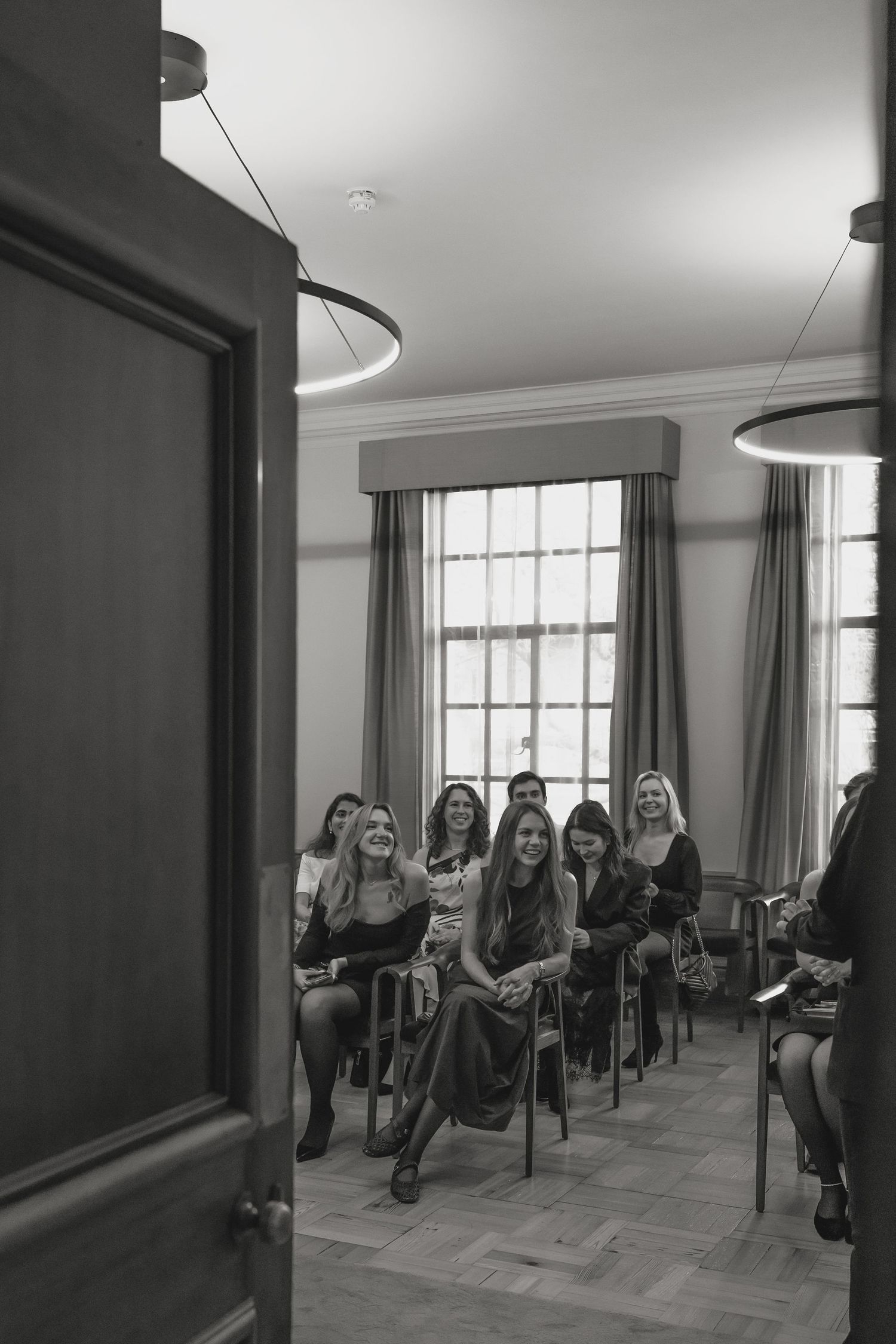 Wedding guests seated in rows in an elegant room with large windows and circular lighting.