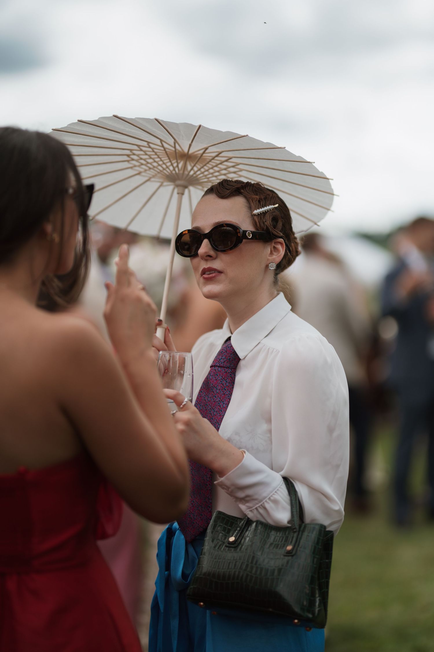 A stylish woman in sunglasses, white shirt, purple tie, holds a parasol and green handbag at an outdoor event.