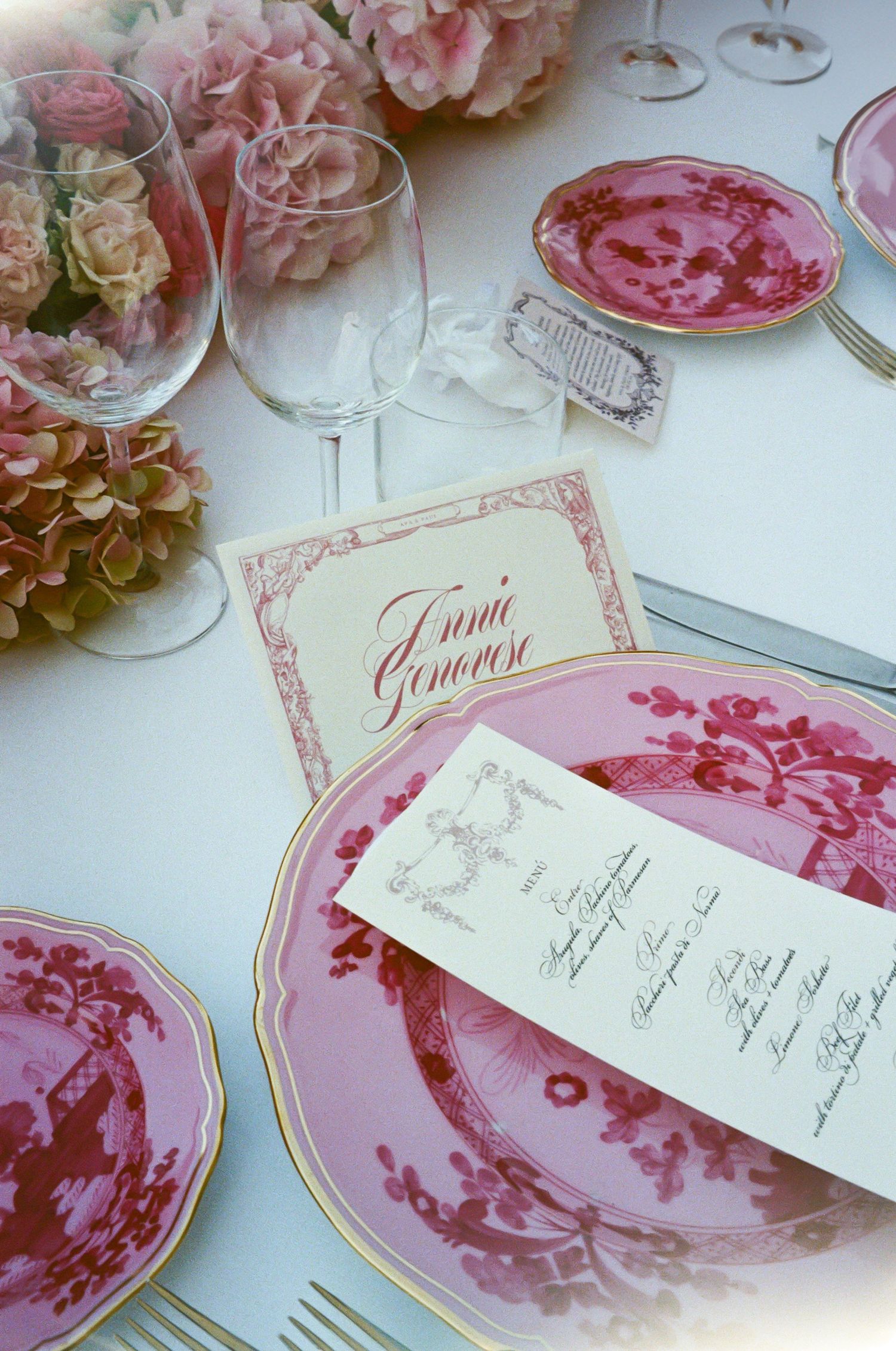 Elegant pink floral table setting with ornate red and white china plates, crystal wine glasses, and a decorative menu card.