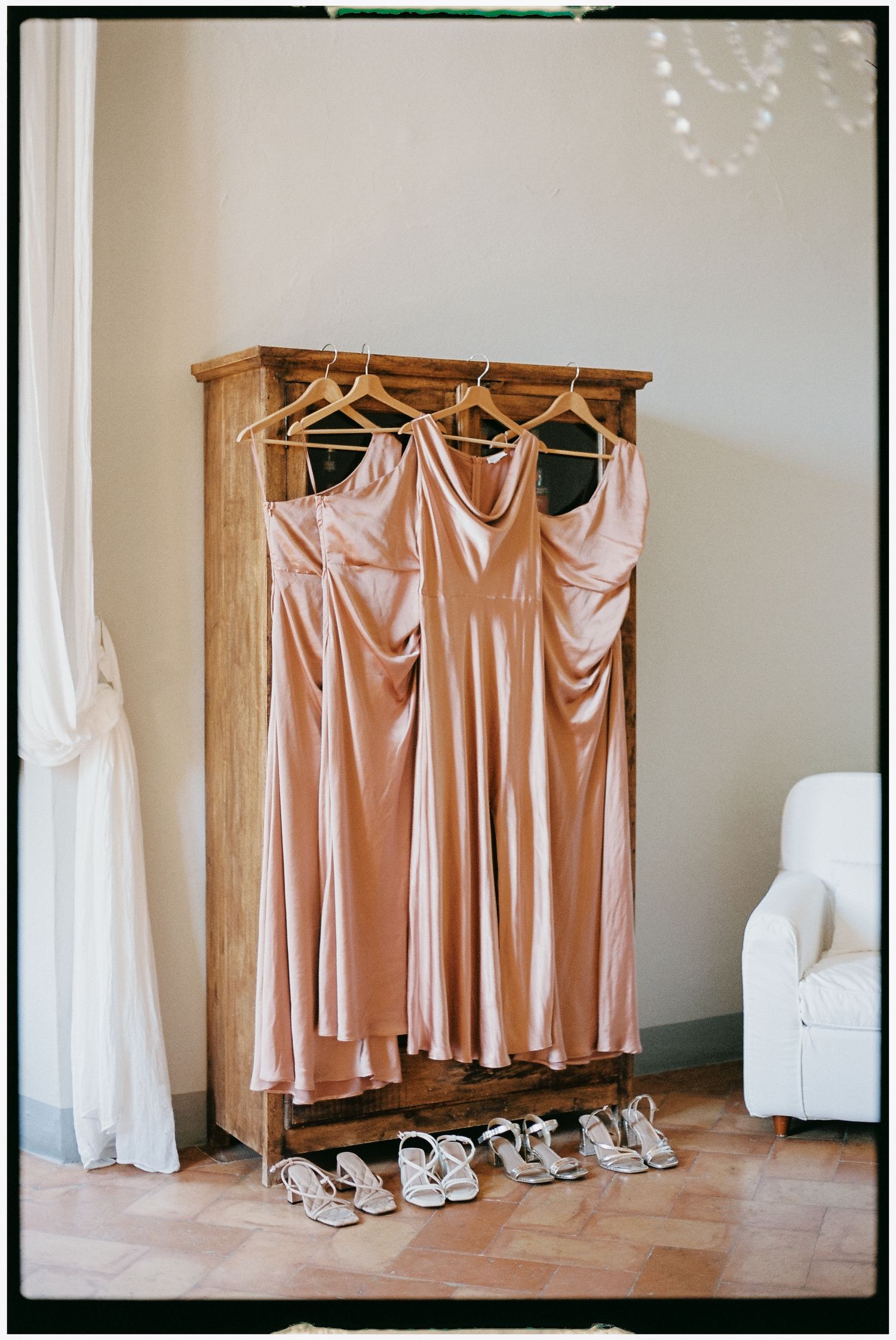 Three blush pink satin bridesmaid dresses hanging on wooden wardrobe with silver heels below.
