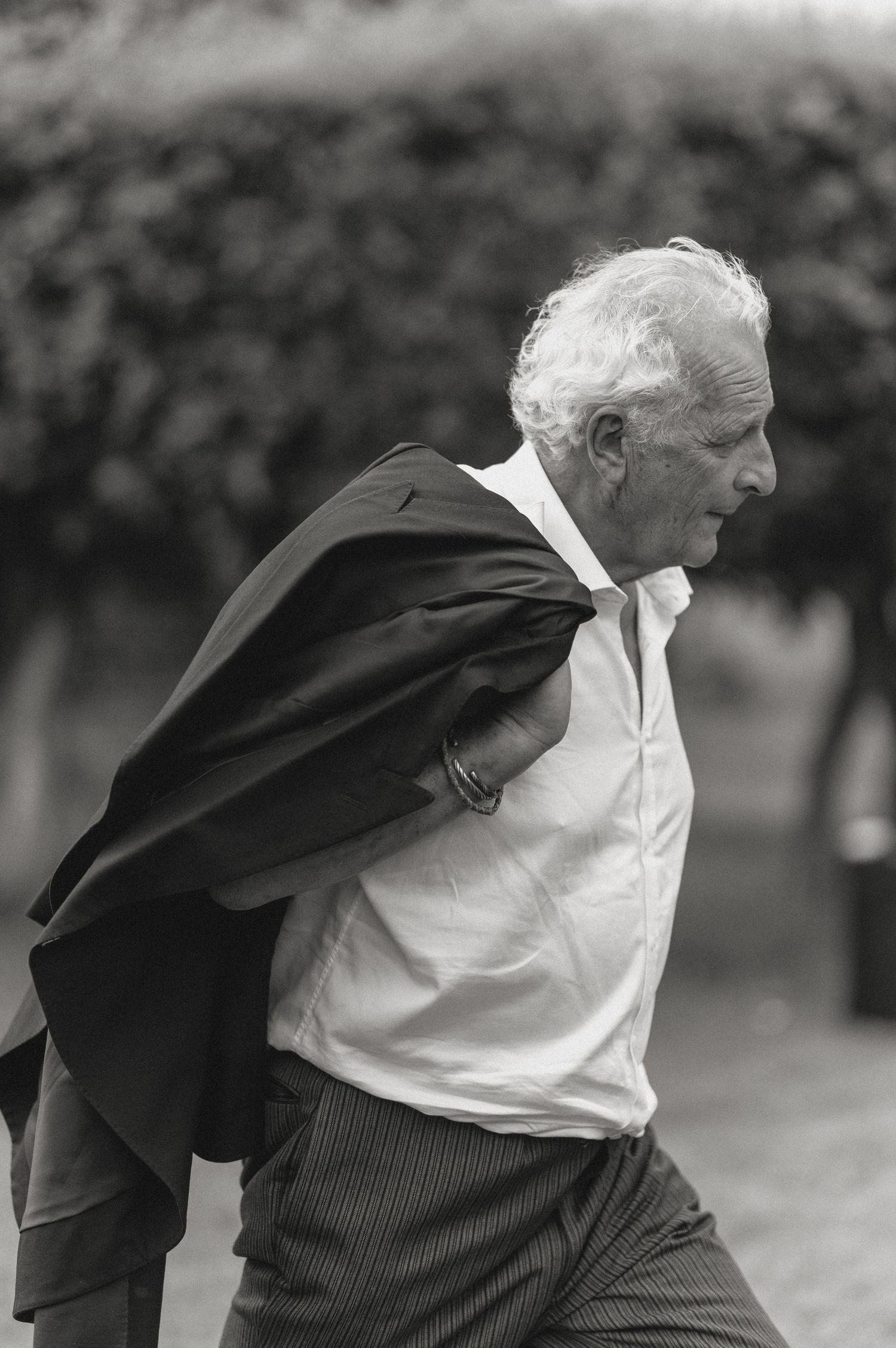 Elderly man with white hair carrying a dark jacket over his shoulder, walking outdoors in black and white photo.