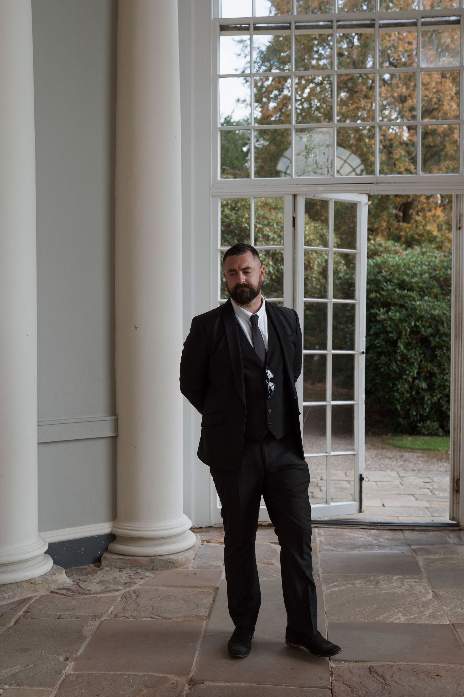 People in formal attire standing in elegant colonnade with large windows.