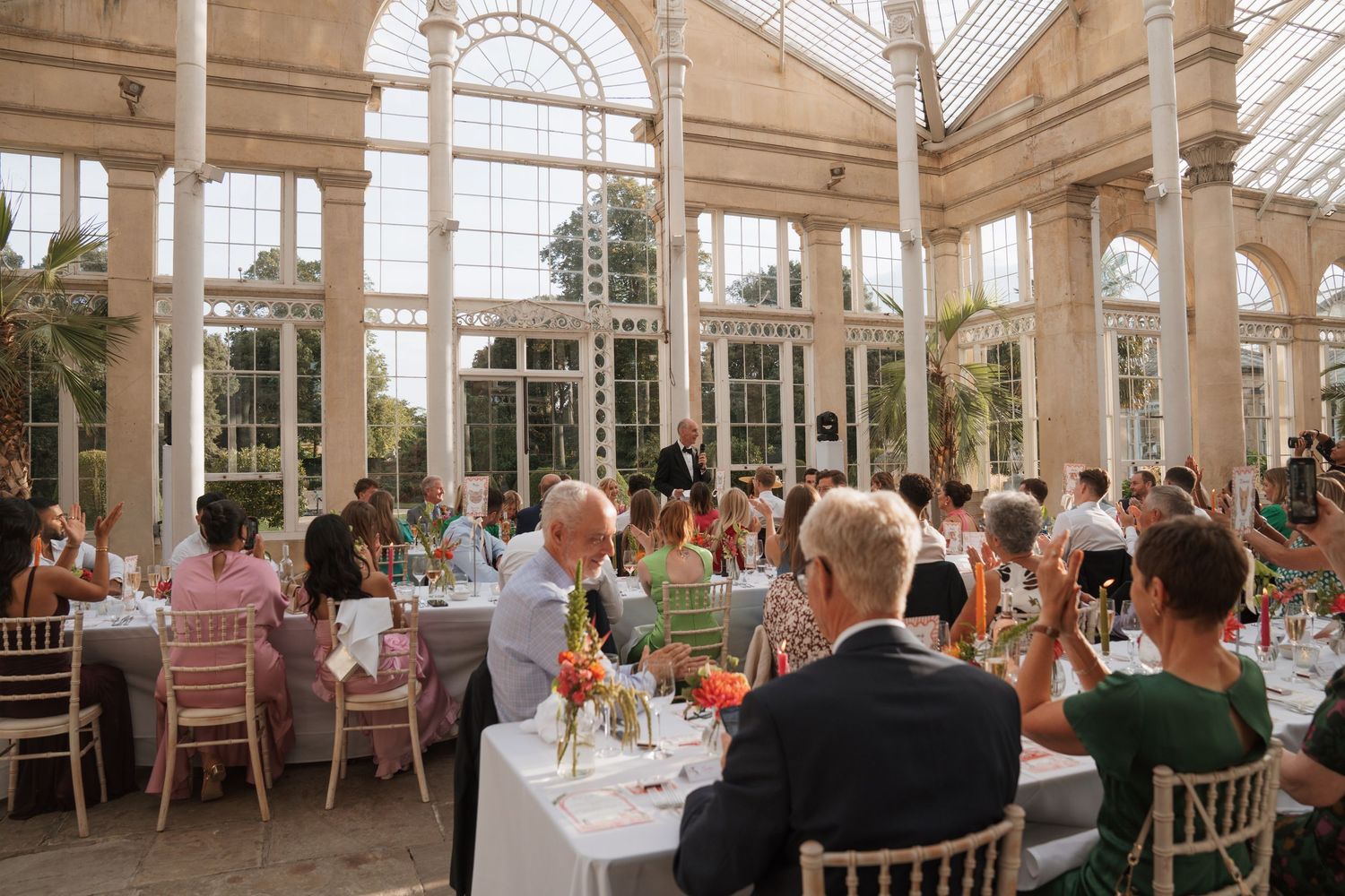 Panoramic view of a sunlit conservatory wedding venue with guests seated at round tables.