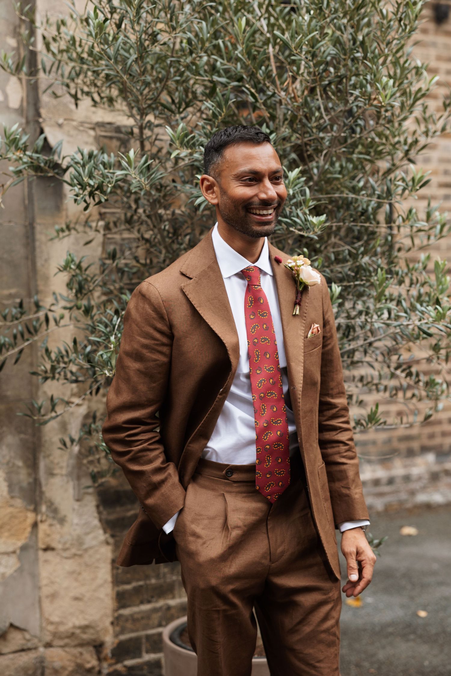 Smiling groom in a brown linen suit, red patterned tie and boutonniere stands beside an olive tree against a brick wall.