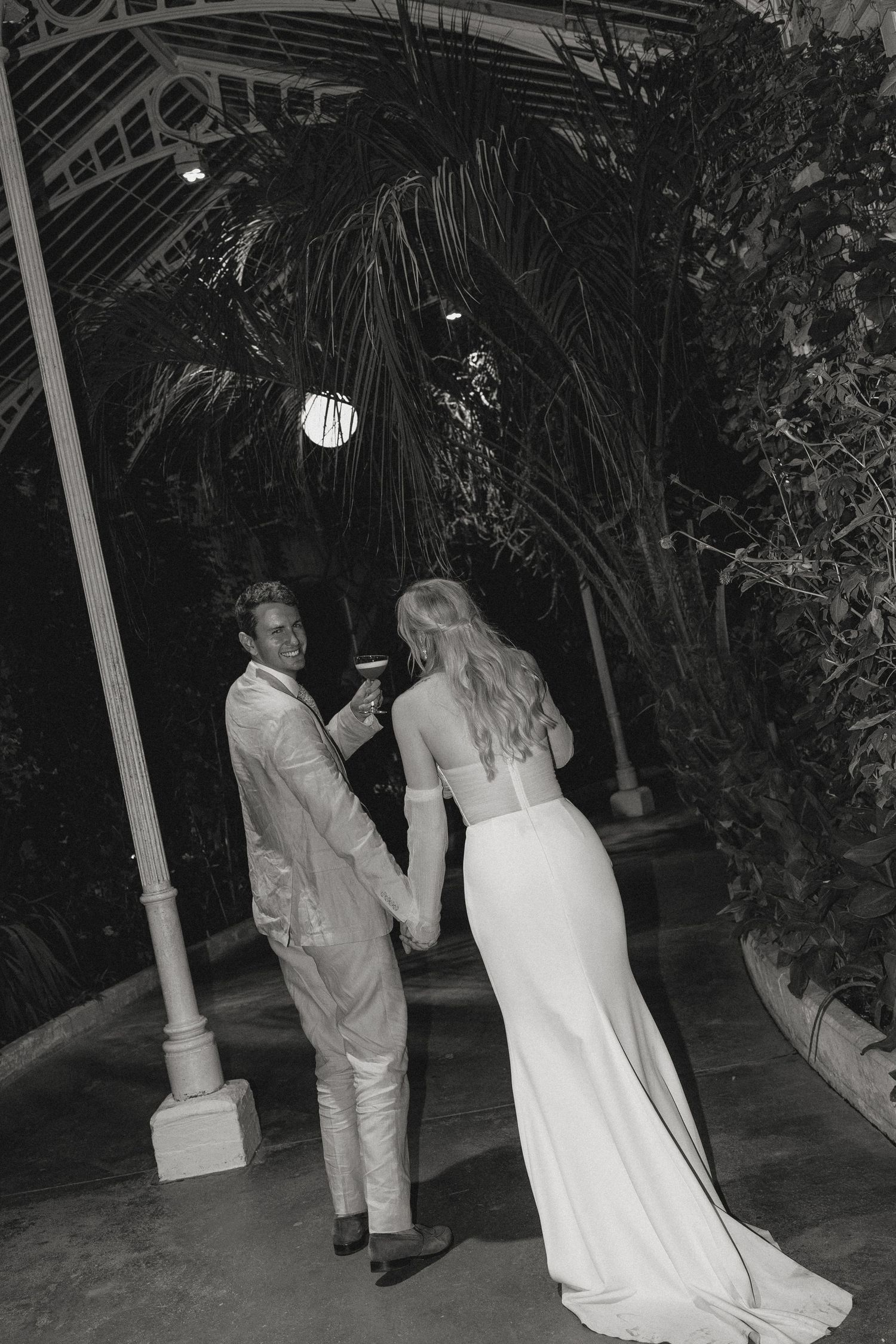 Black and white photo of a bride and groom holding hands and toasting outdoors at night surrounded by tropical greenery.