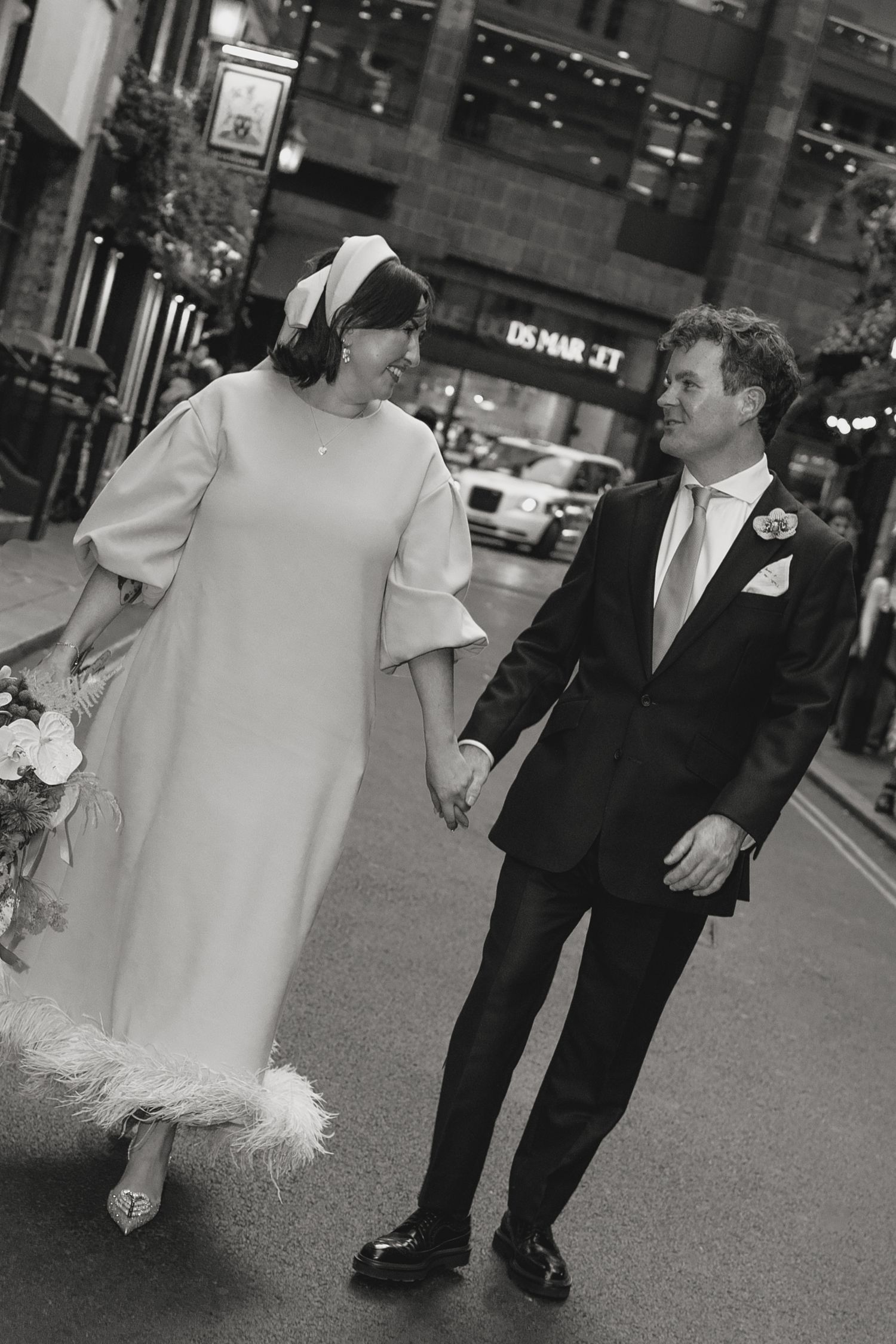 Black and white photo of a bride and groom holding hands, walking down a city street on their wedding day.