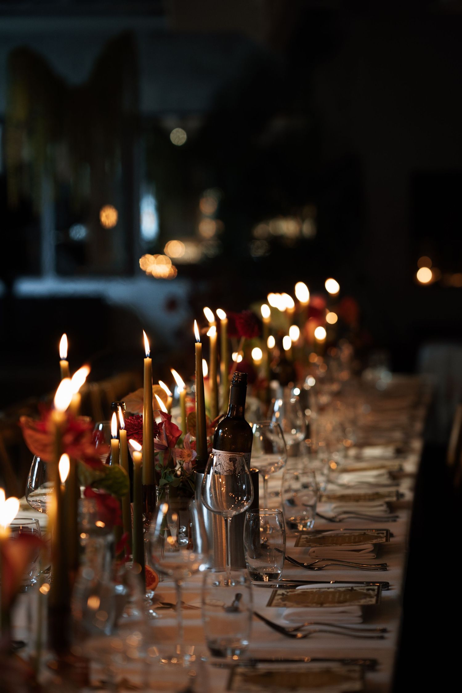 Long dining table set with glowing candles and place settings in dark ambient lighting.