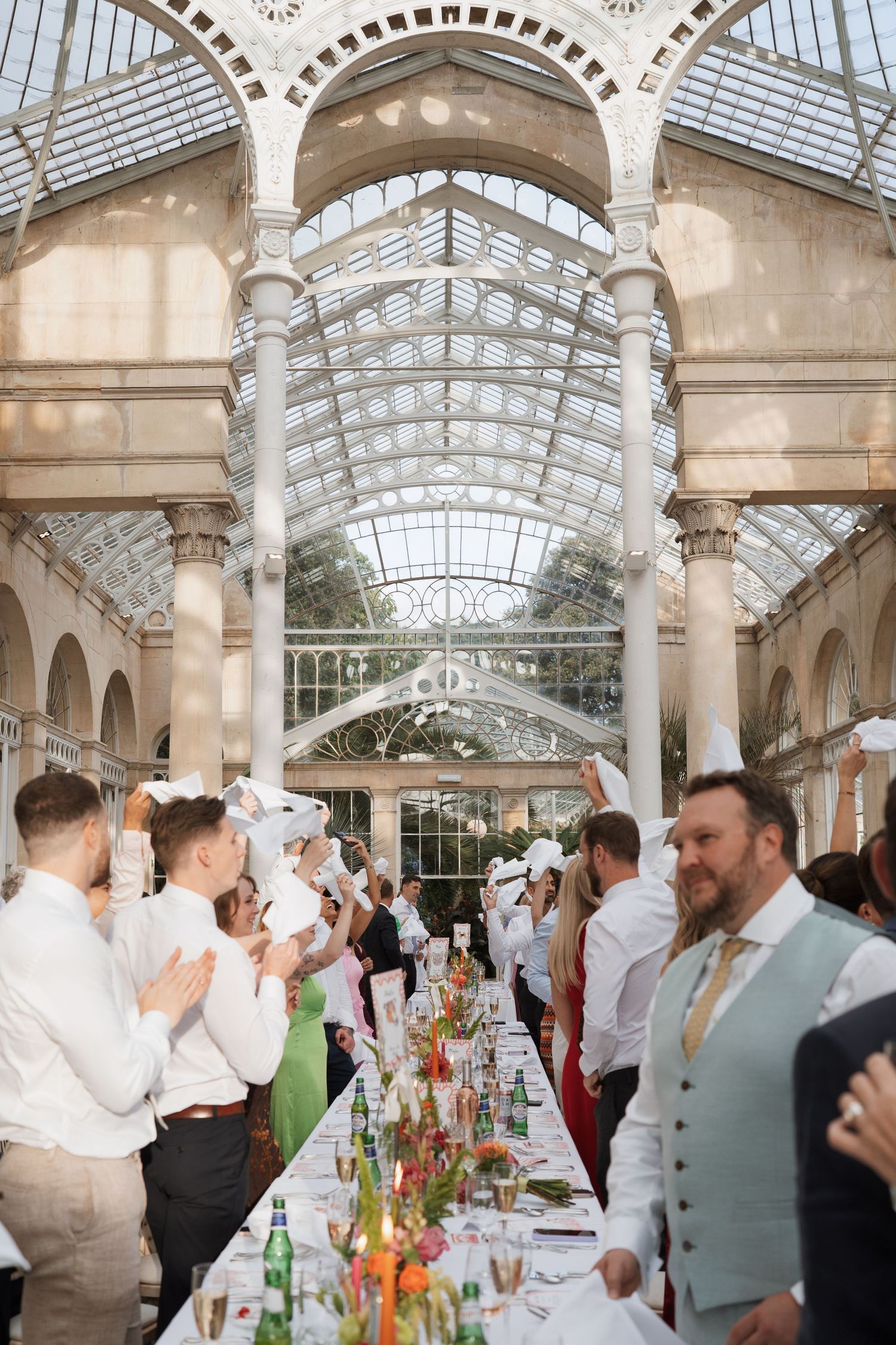 Wedding guests celebrate at an elegant indoor reception venue with ornate glass ceiling and architectural details.