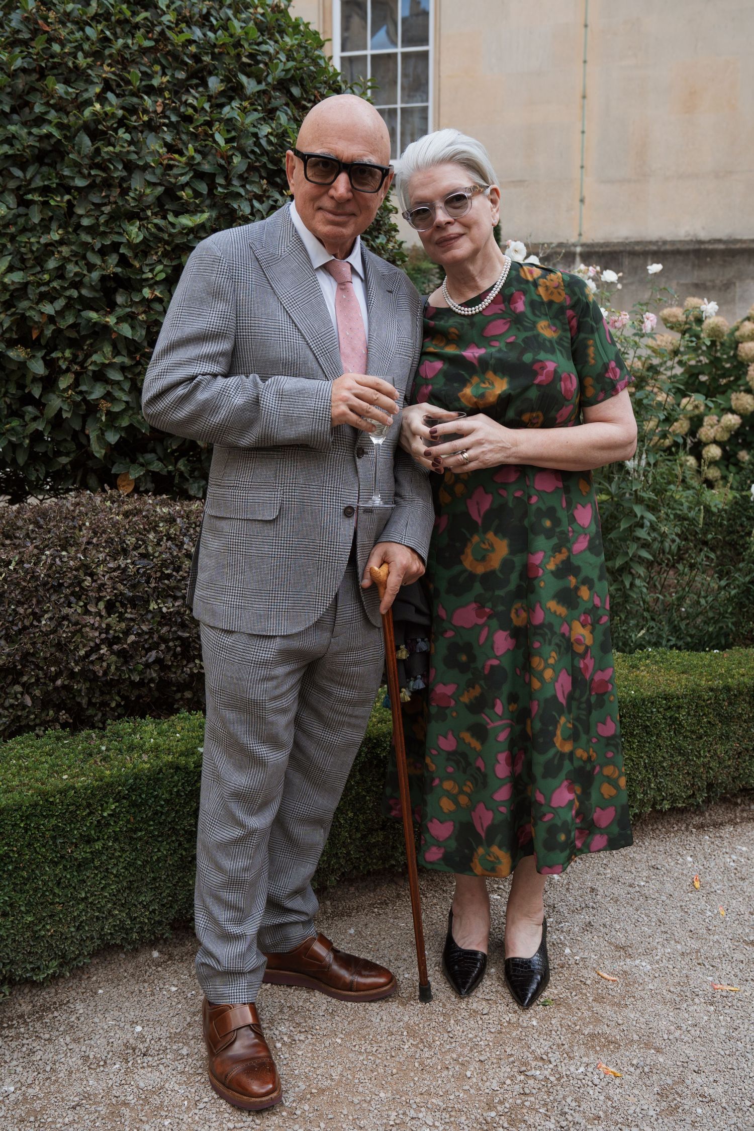 Elegant couple posing in garden, man in grey suit with cane, woman in floral green dress, outdoor event.