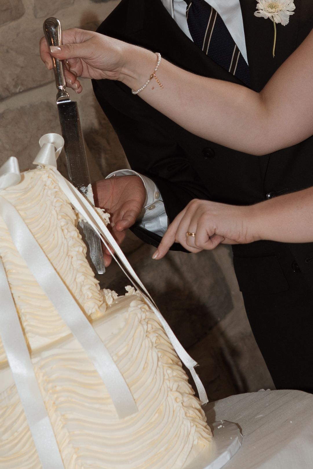 Hands cutting into an elegant white wedding cake with decorative frosting patterns.