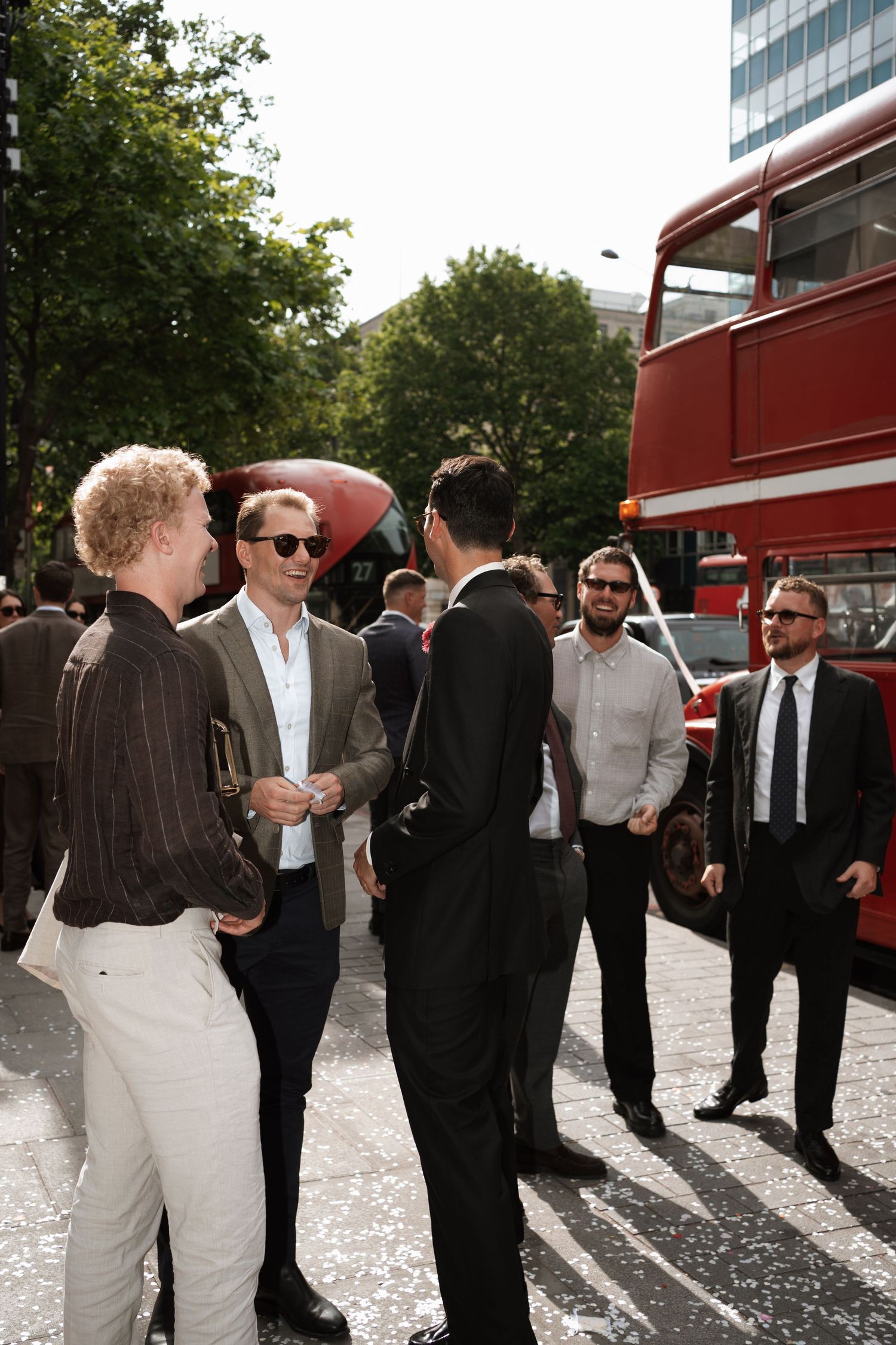 Wedding guests in formal attire socializing outdoors near red buses.