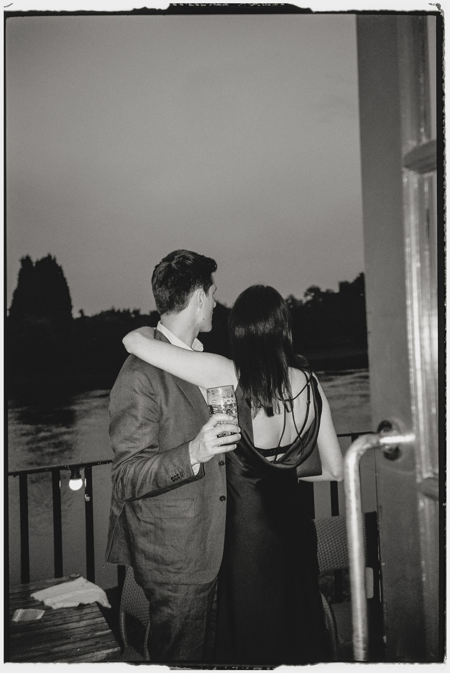 Black and white photo of a couple embracing on a waterfront deck at dusk, man holding a drink.