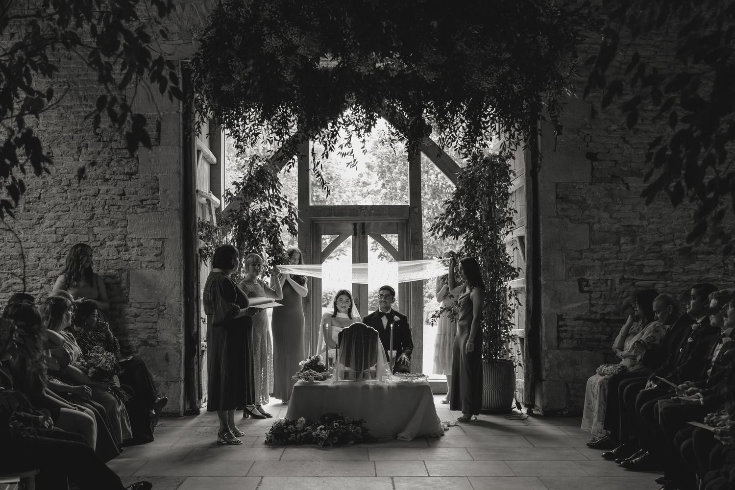 Black and white wedding ceremony photo in rustic barn venue with floral arch, guests seated, couple at altar.