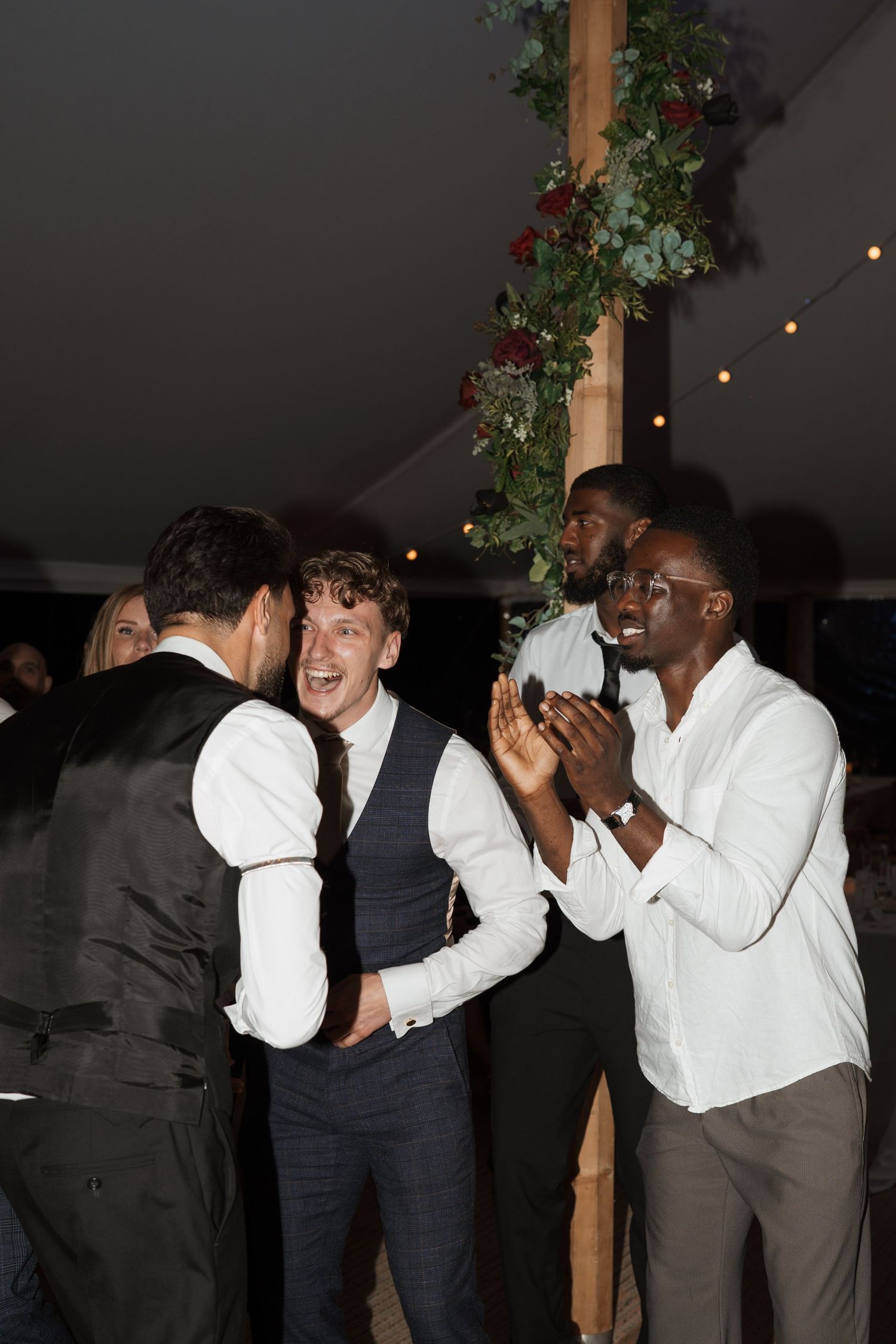 Wedding party members share laughs and celebrations under decorative tent lighting.