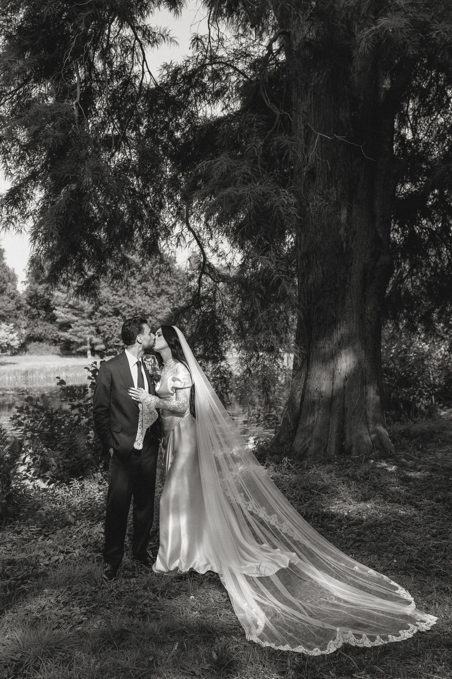 A wedding couple poses under a large tree with the bride's long veil flowing behind them.