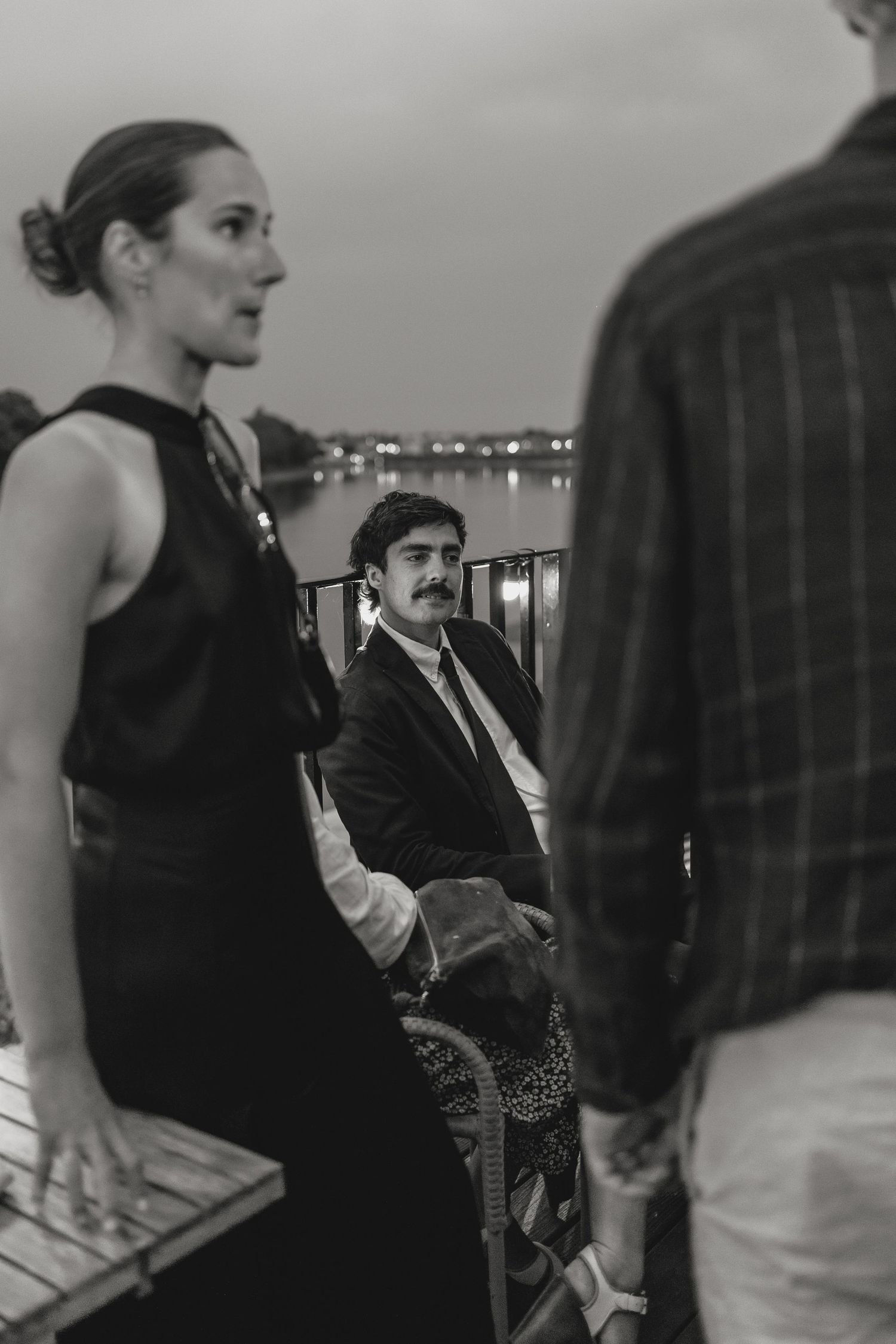 Evening wedding reception guests gather on outdoor deck with city lights in background.