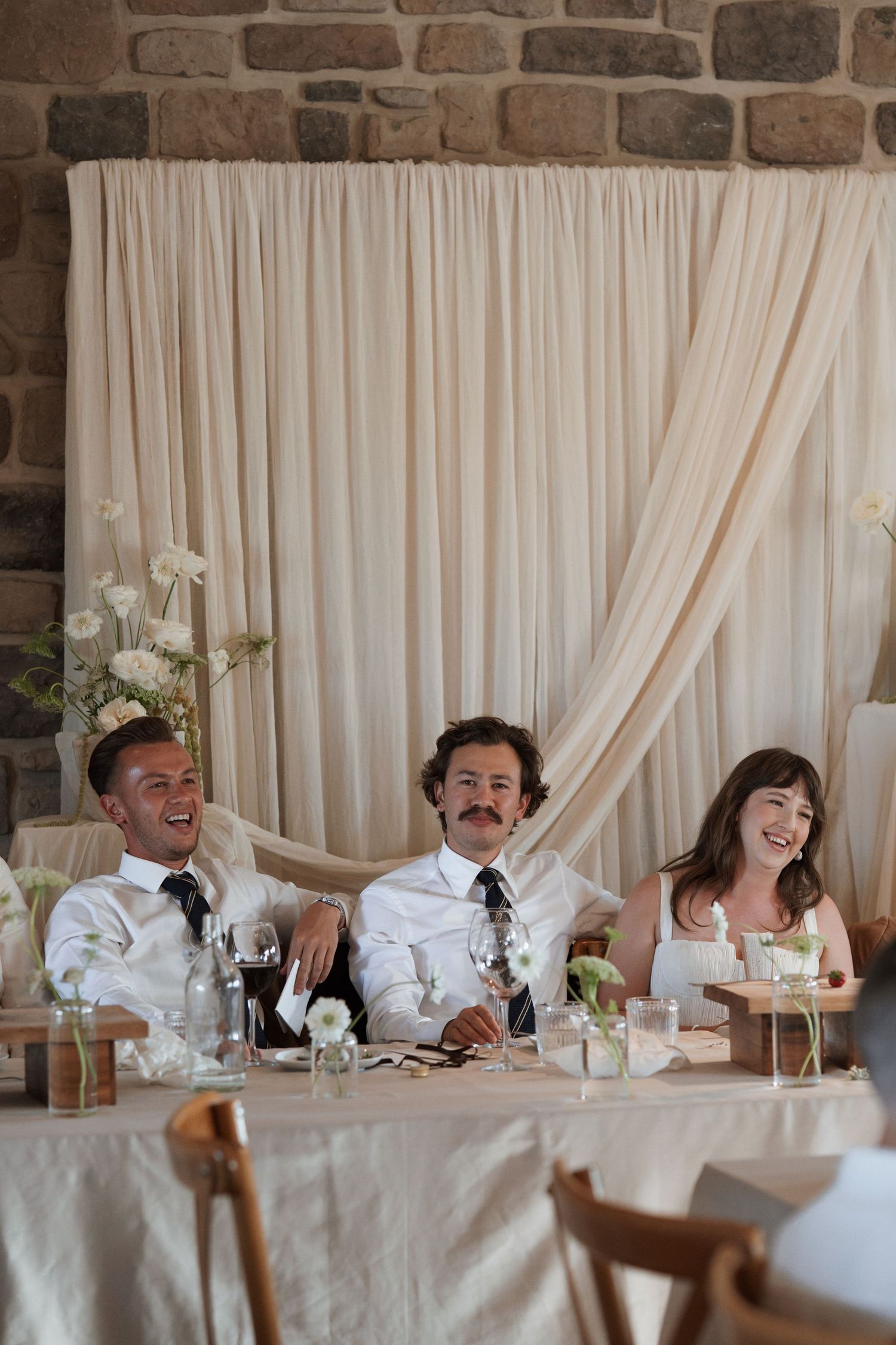 Wedding party enjoying laughter and conversation at the head table with white draping backdrop.