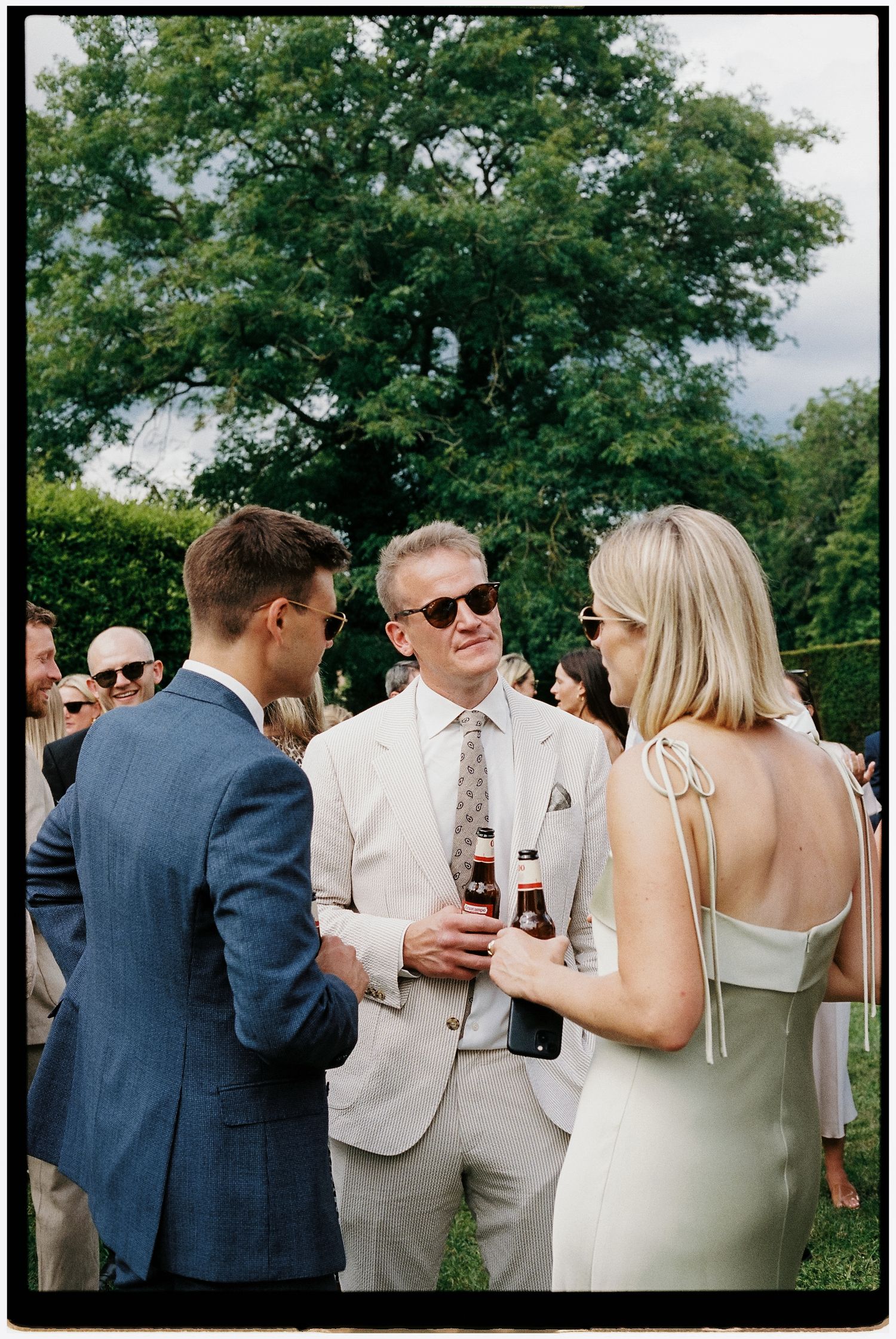 Guests in elegant attire socializing outdoors at a summer wedding reception, holding drinks among lush greenery.