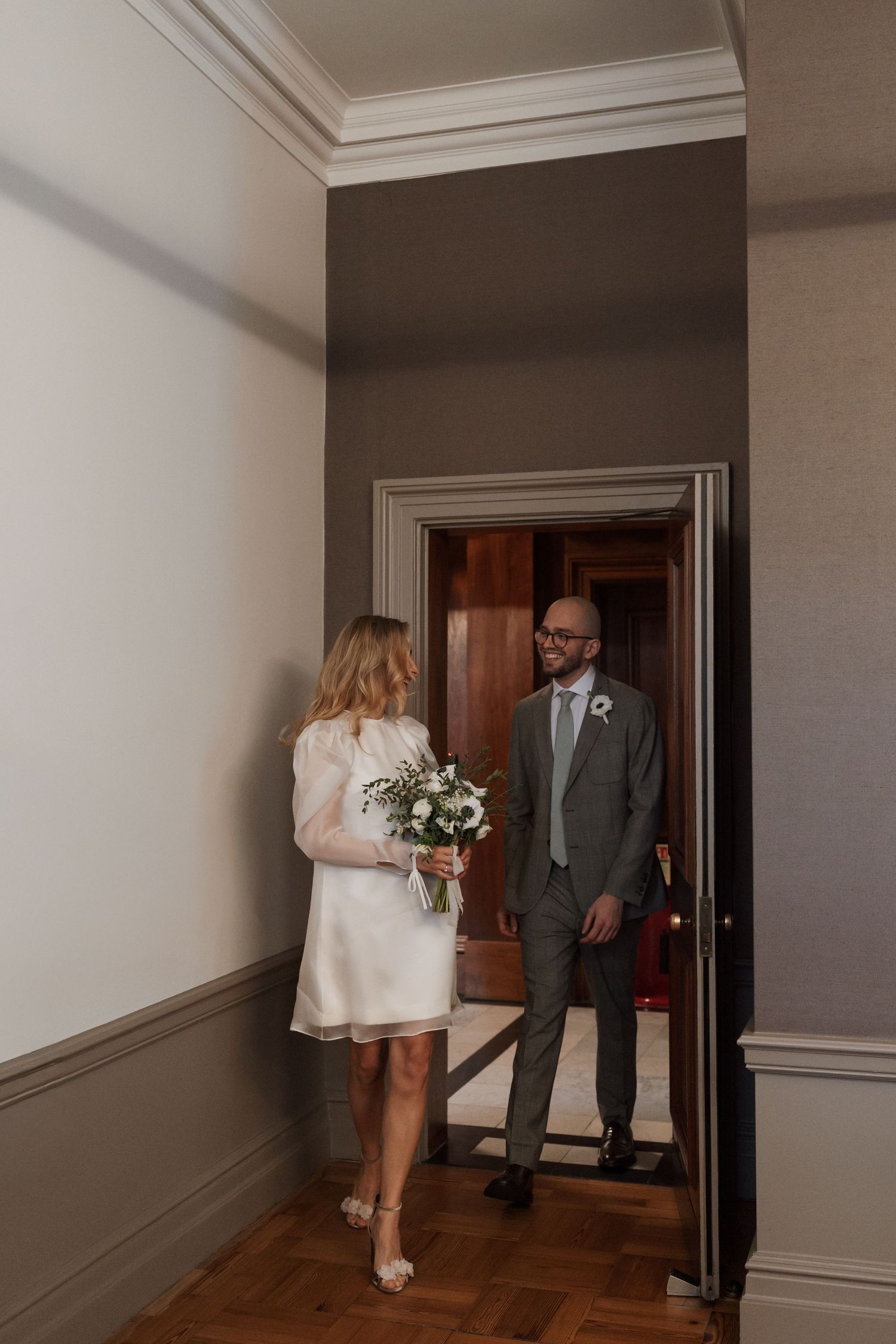 A couple stands in a doorway of an elegant interior with grey walls and wooden floors.