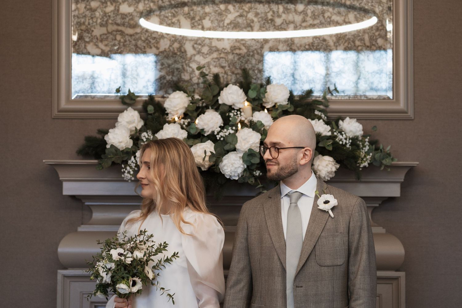 A couple shares a tender moment in front of an elegant white floral arrangement and ornate windows.