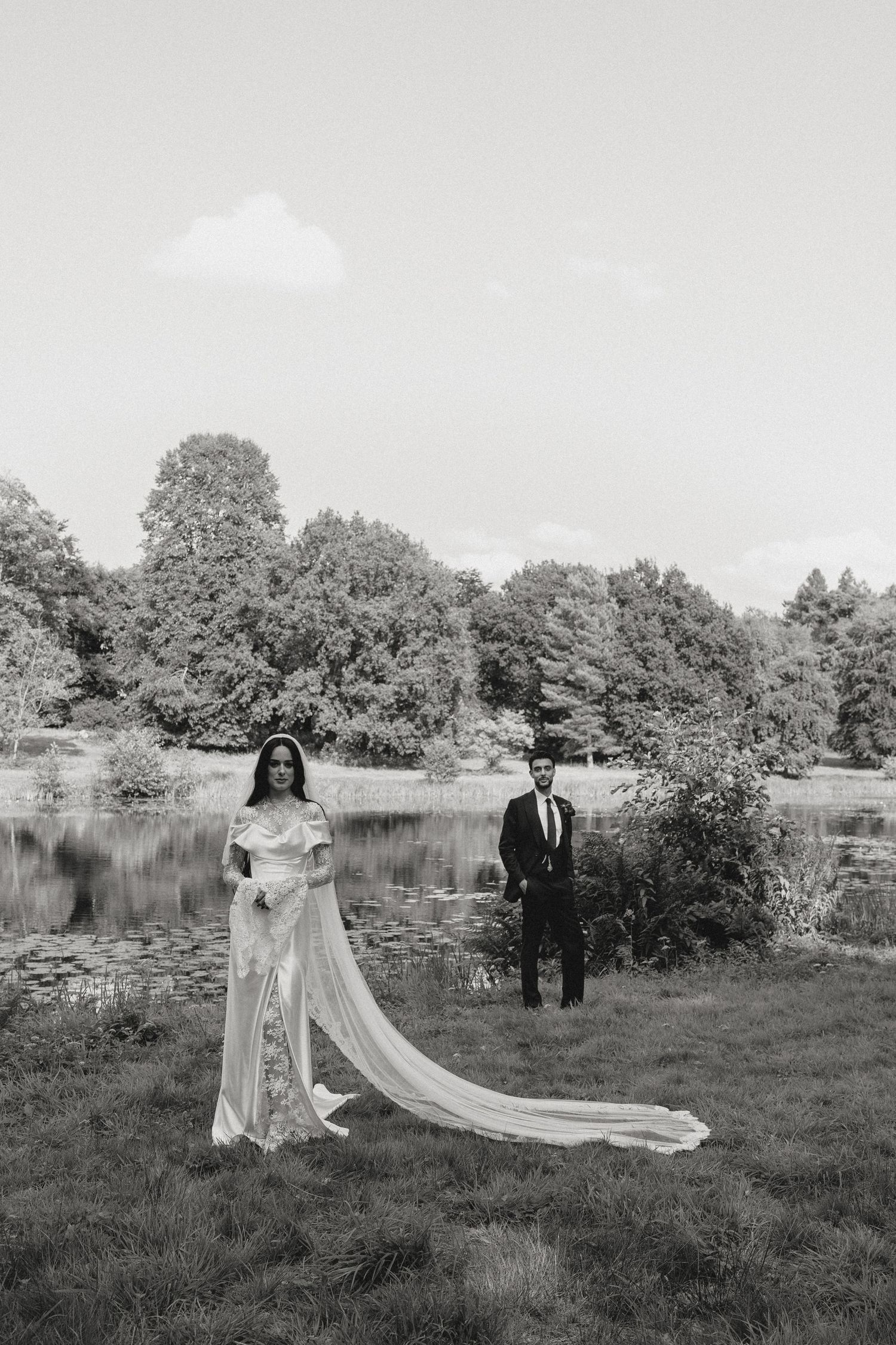 A couple poses in wedding attire near a serene lake surrounded by trees in a black and white landscape.