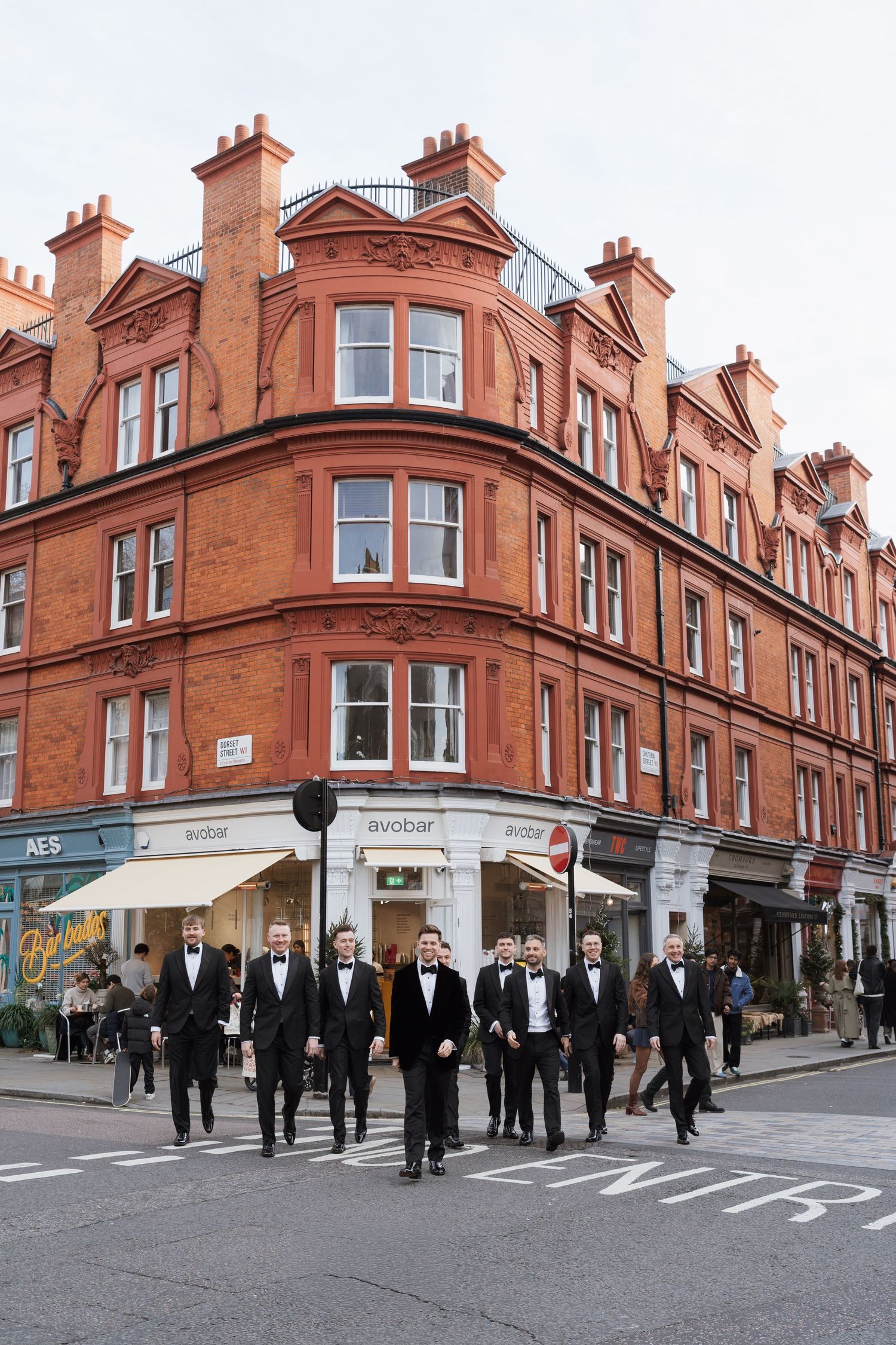 Wedding party in formal attire walks along red brick buildings in urban setting.