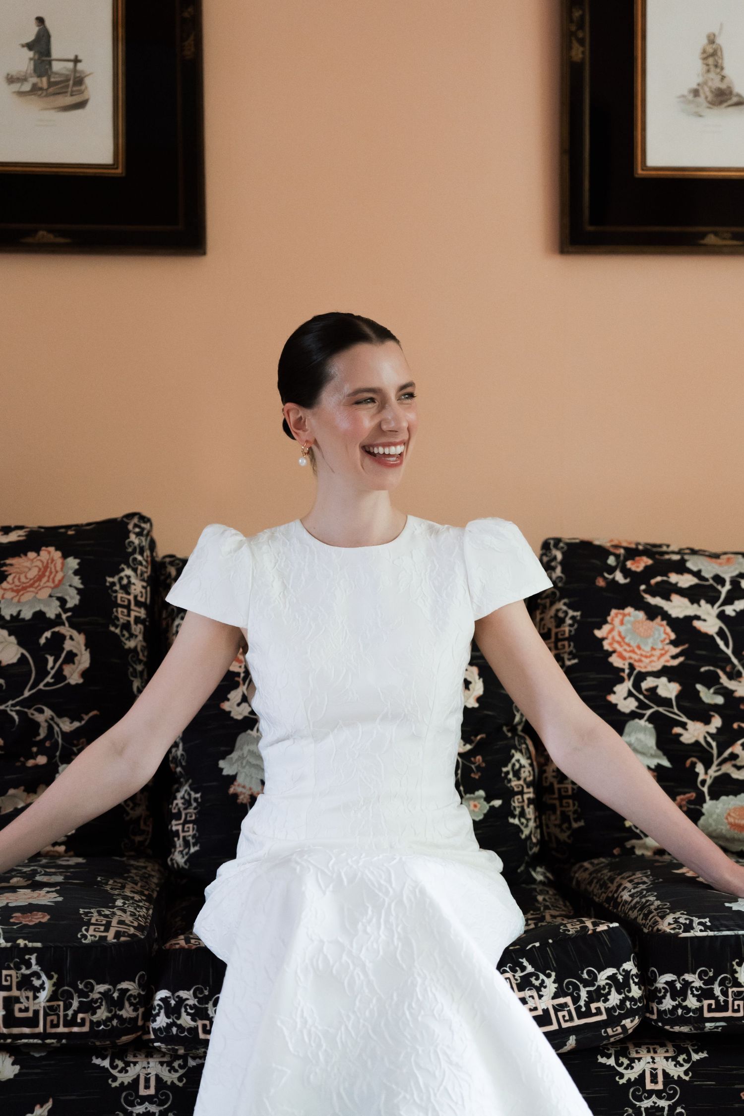 Smiling woman in a white puff-sleeve dress with textured skirt sits on a floral black sofa in front of a peach wall with framed prints.