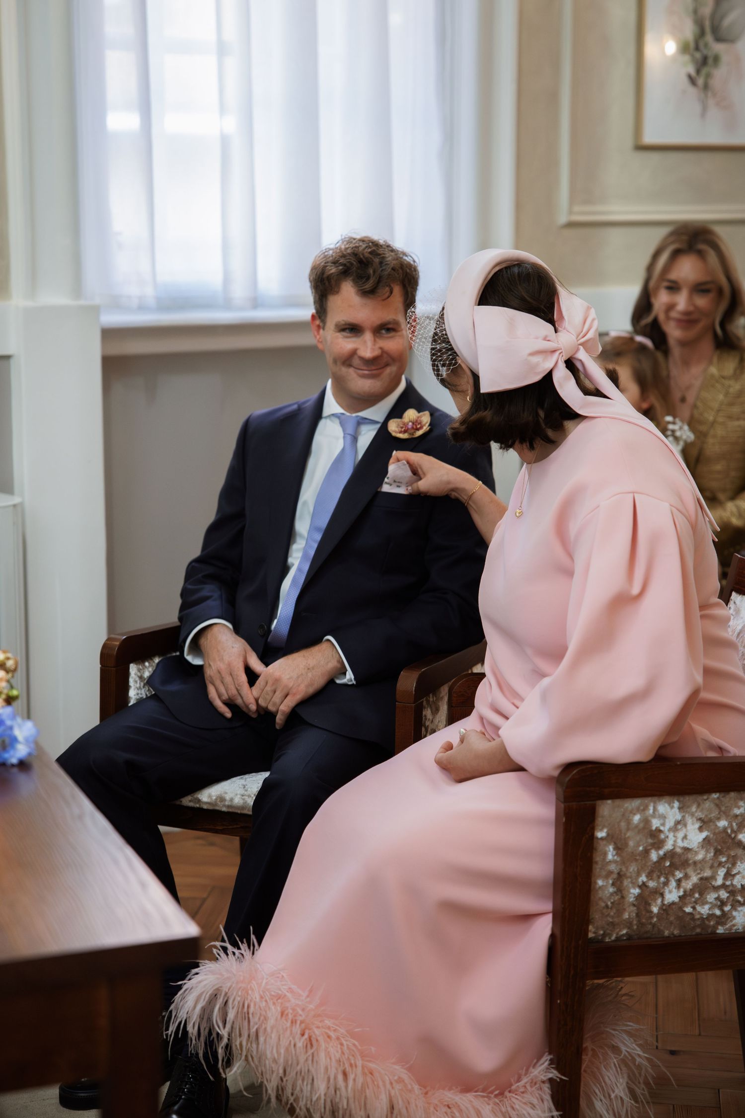 A couple sits together at a formal event, with the woman wearing a pink dress with feather trim.