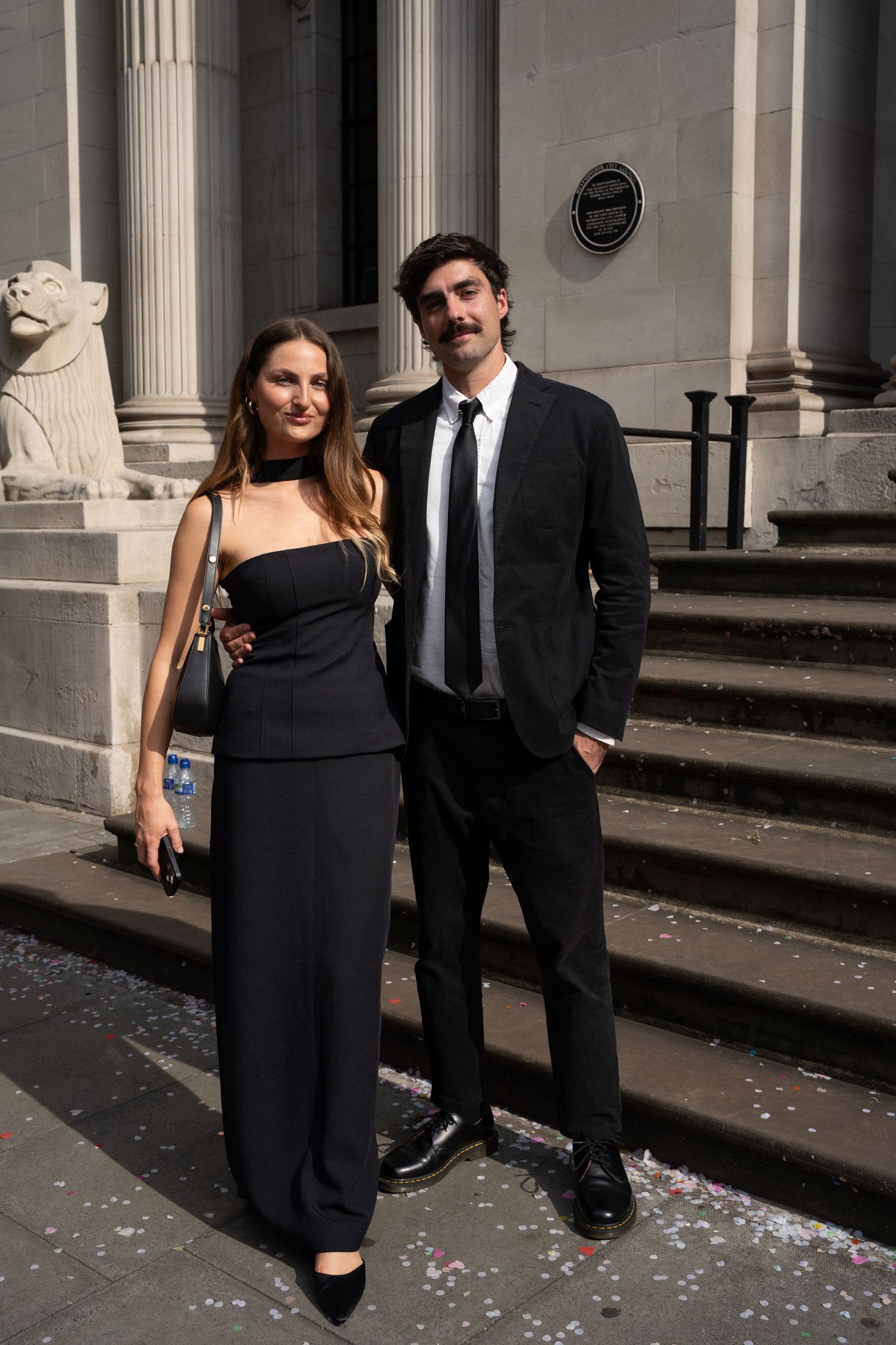 Elegant couple in formal attire walking outside a classical building with columns.